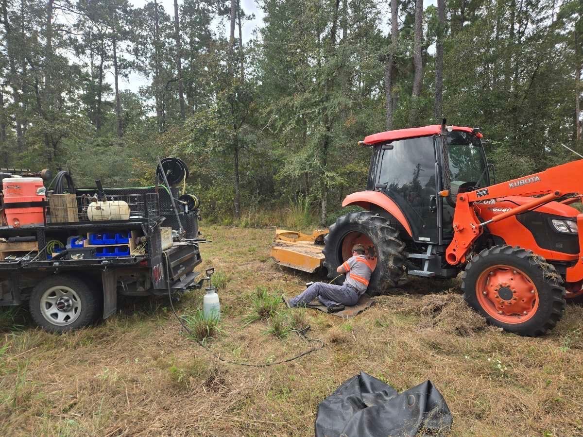 A person works on a tractor near a truck in a field, trees in the background.