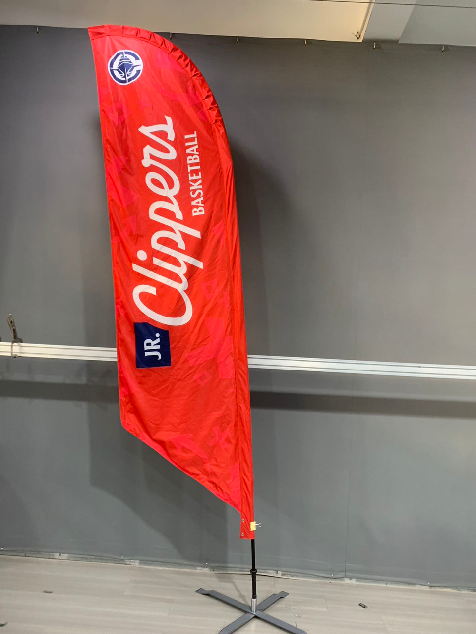 Red Clippers flag with logo and text against a gray wall, held up by a black pole and a silver base.