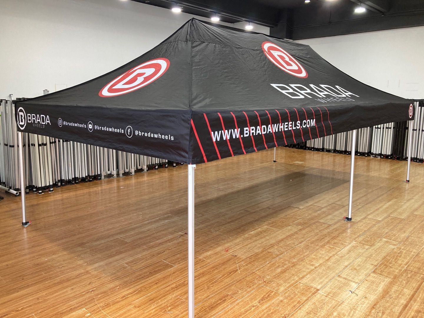 Black branded canopy tent with white logos and website, set up indoors on a wood floor.