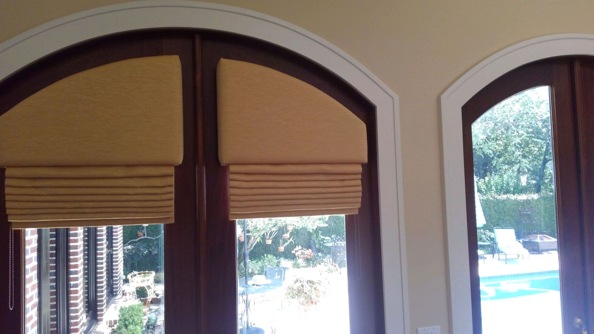 Arched windows with gold Roman shades; wooden doors with view of a pool.