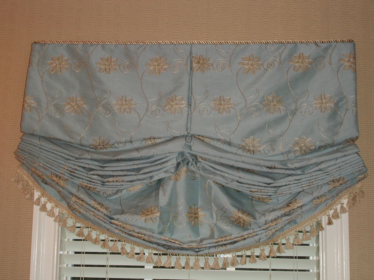 Light blue, floral embroidered window valance with gold trim and tassels over a window.