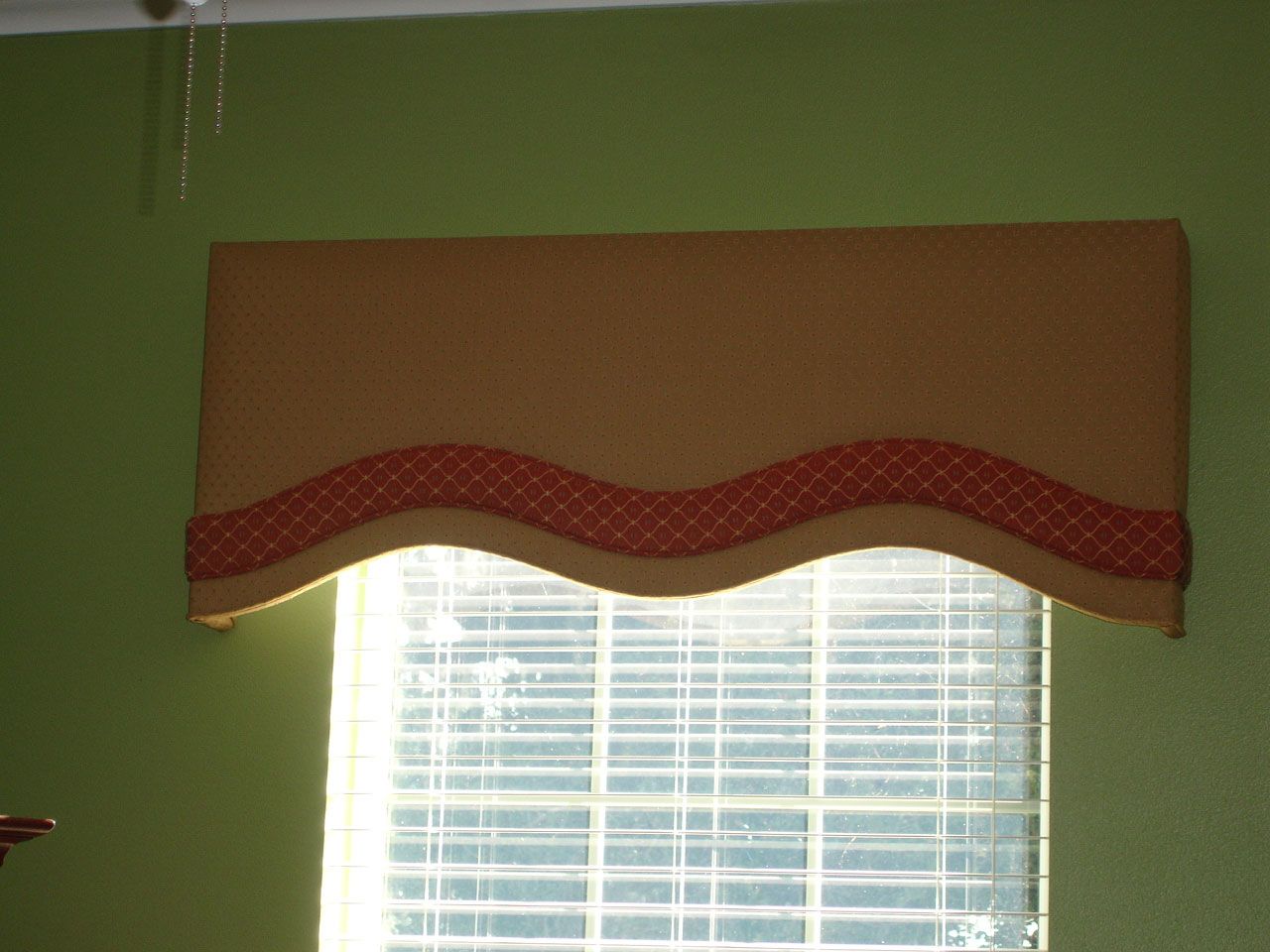 Tan and red scalloped valance over a window with blinds on a green wall.