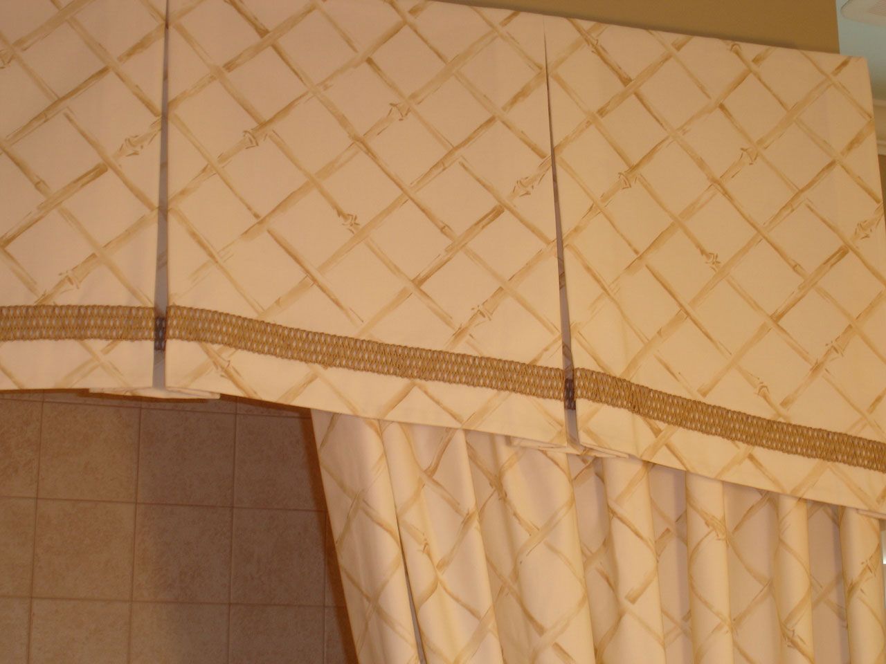Cream-colored fabric valance and curtain with brown diamond pattern and trim, hanging in front of tiled wall.