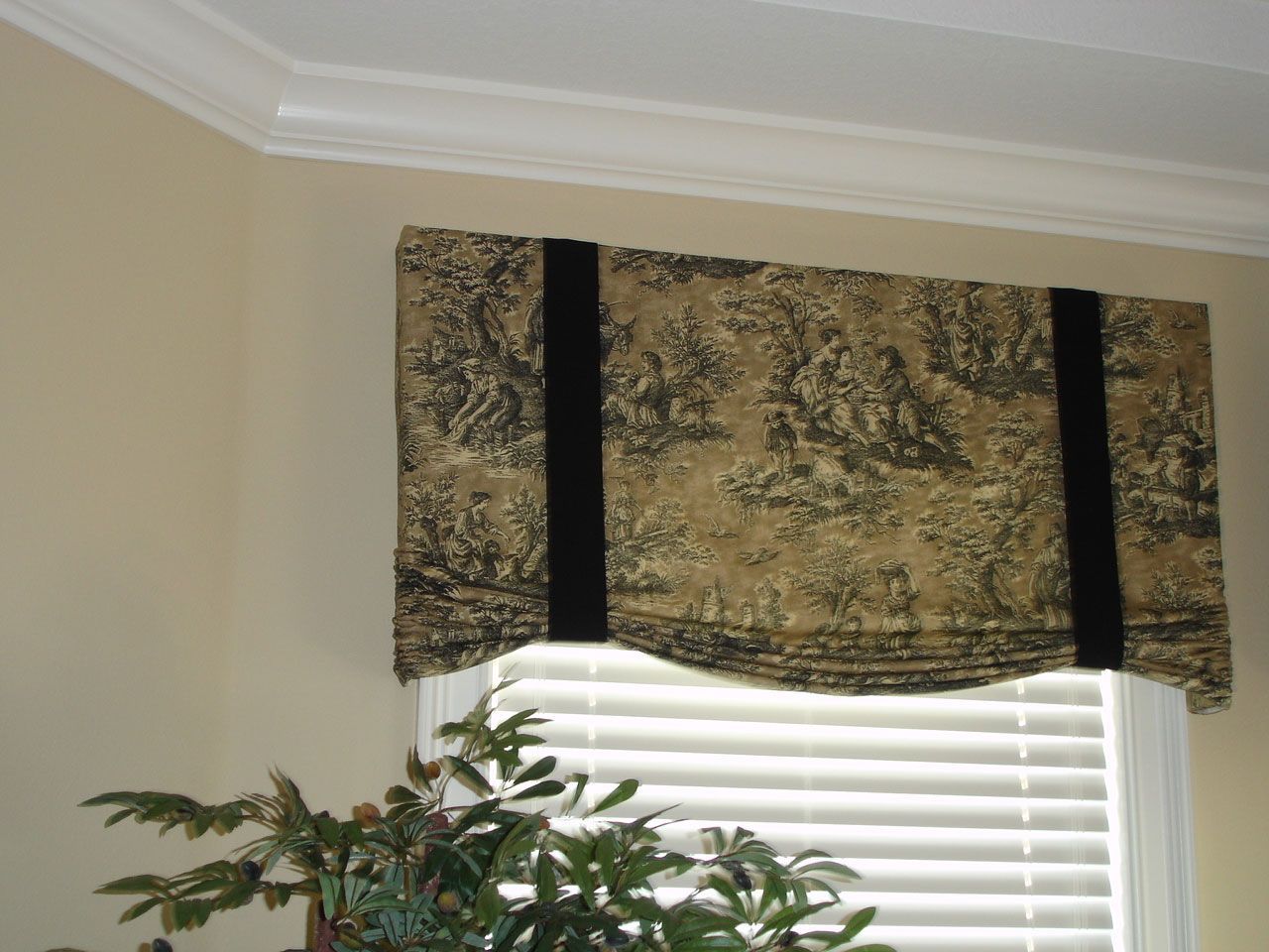 Beige and black patterned window valance over a window with closed white blinds.