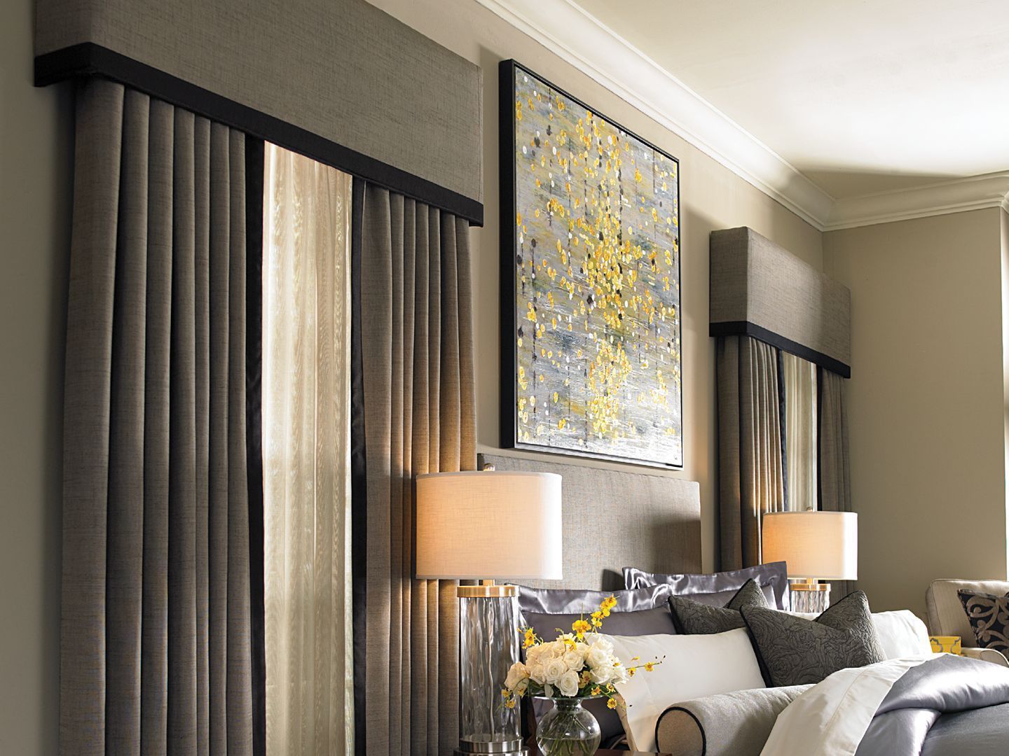 Bedroom with gray curtains, painting with gold accents, lamps, and a bed with decorative pillows.