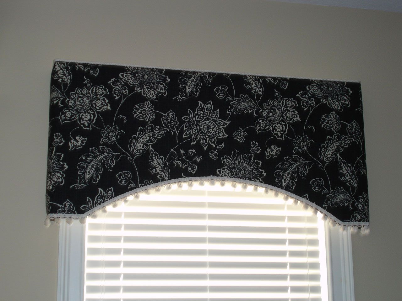 Black and white floral fabric valance with white pom-pom trim over a window with white blinds.