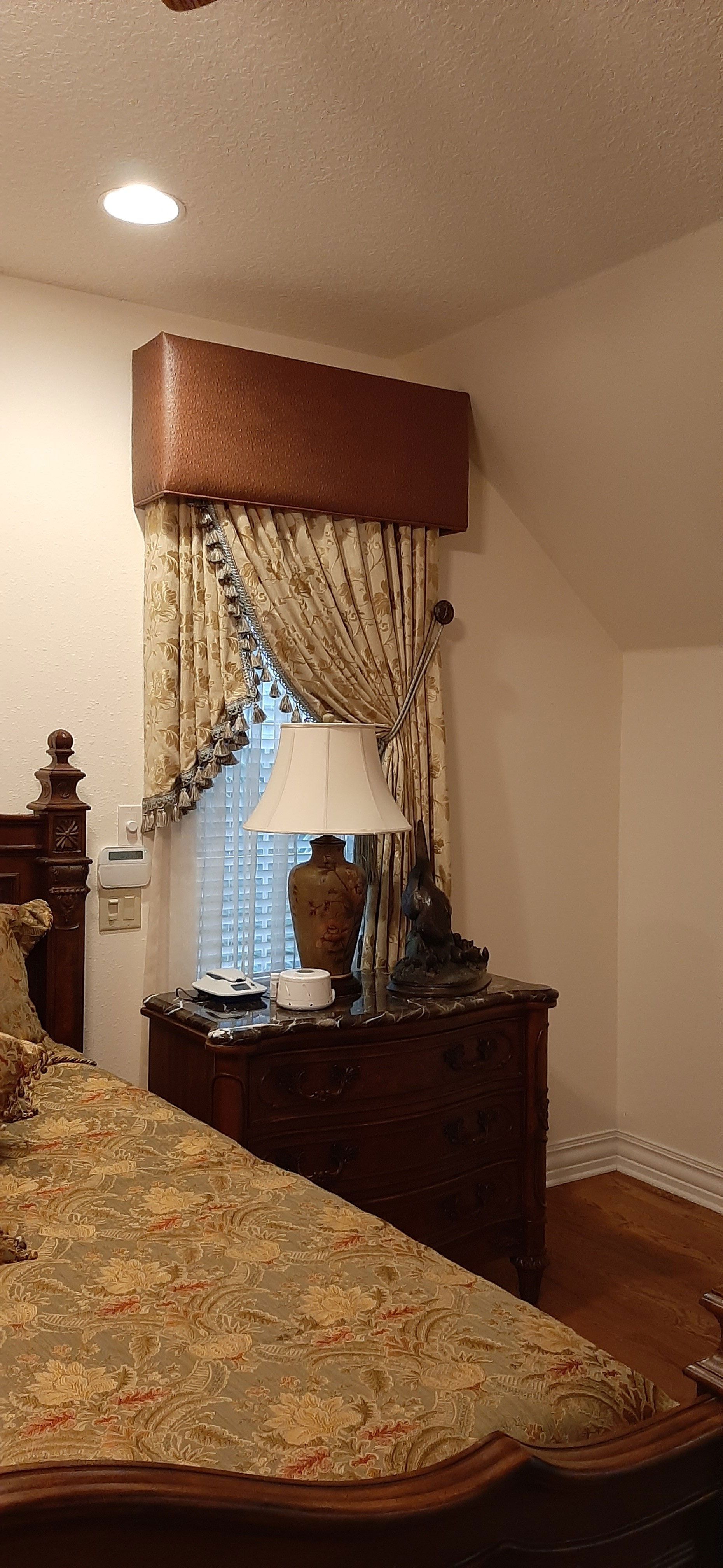 Bedroom with floral bedspread, ornate curtains, lamp on nightstand, and brown headboard.