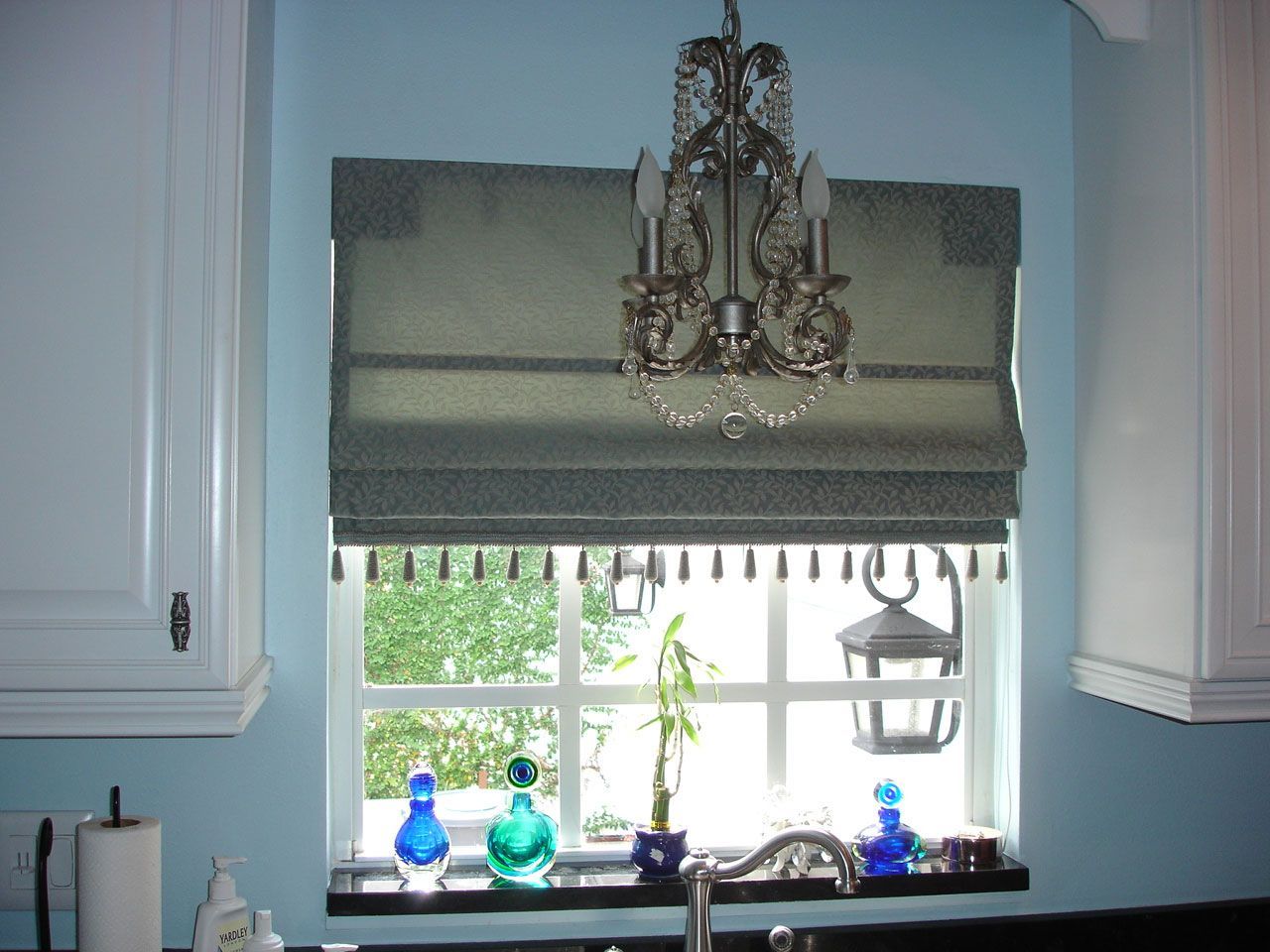 Kitchen window with blue walls, white trim, Roman shade, chandelier, and decorative items on the sill.