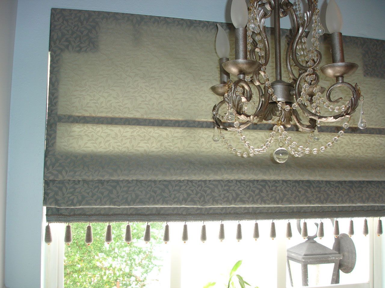 Roman shade with fringe, chandelier, and window. Greenish-blue fabric, decorative details, and a light blue wall.