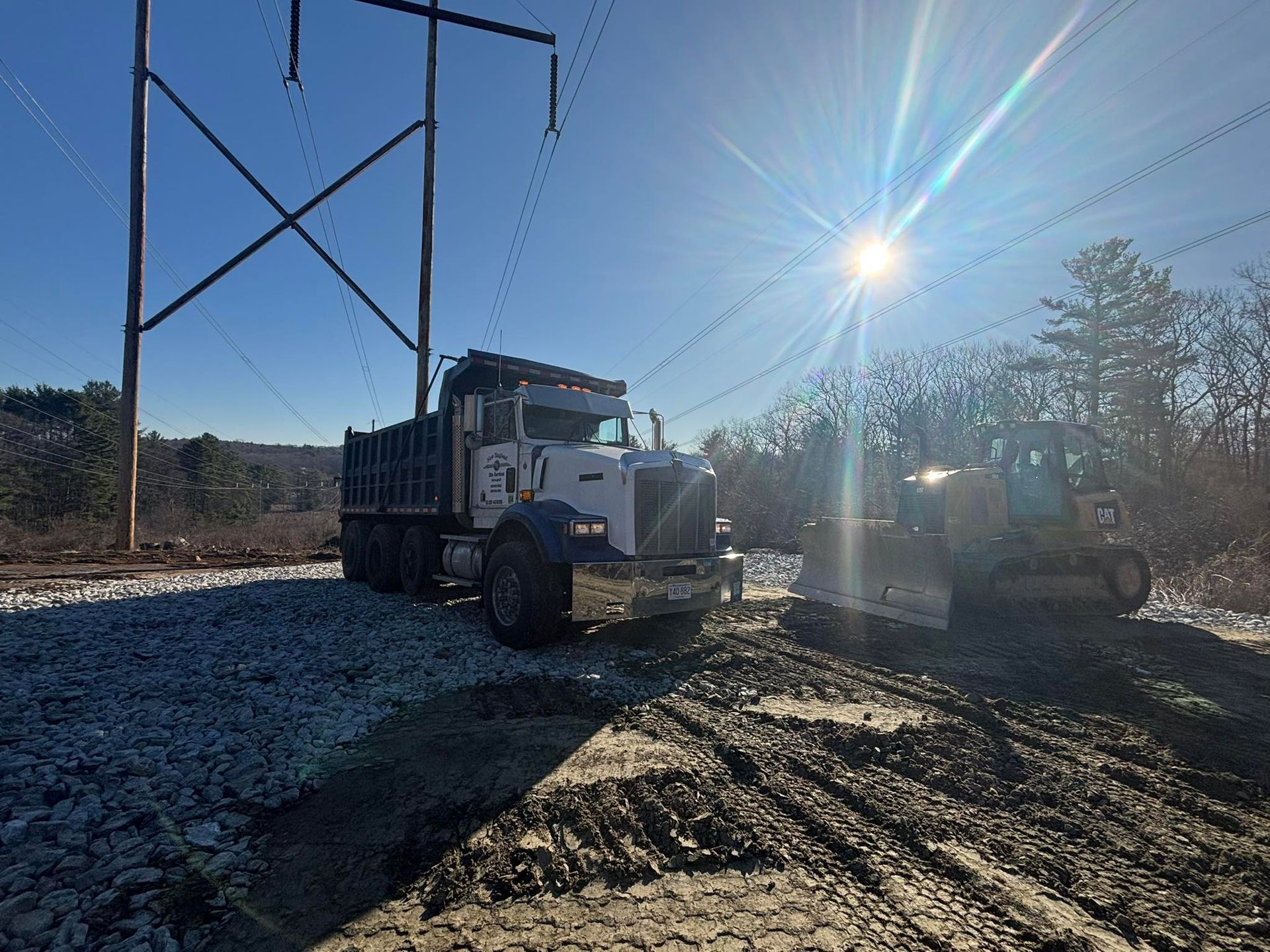 White dump truck on gravel road beside utility poles, with bright sun and a tractor in the background