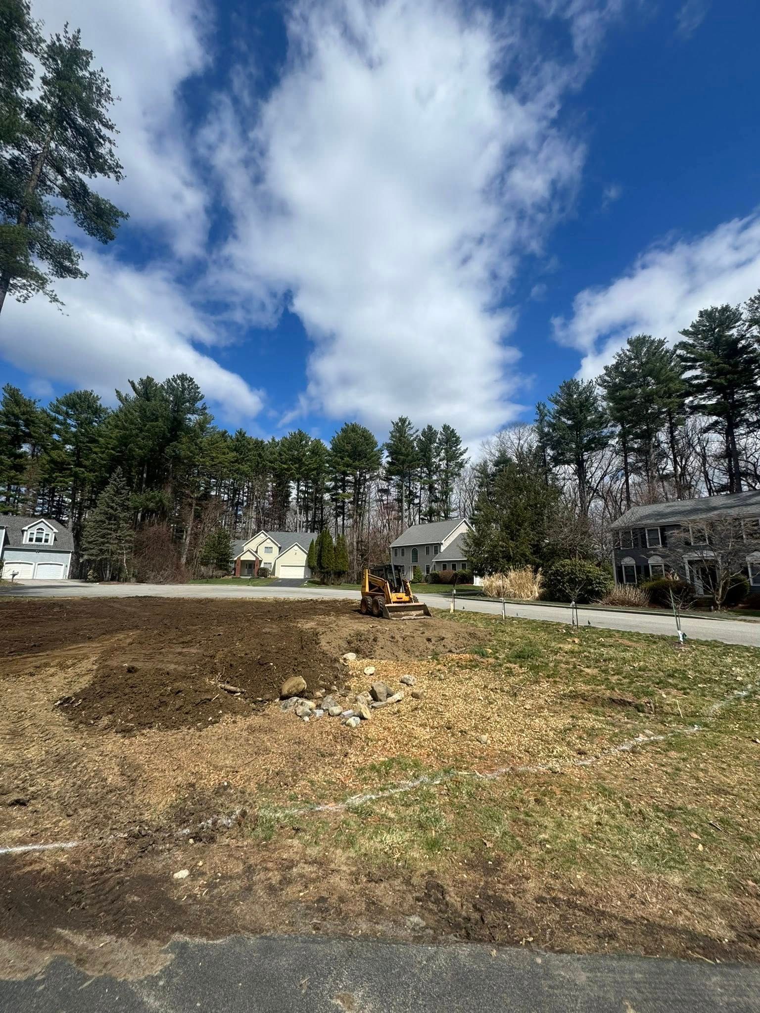 Construction equipment clearing a dirt lot beside a road, with trees and houses under a cloudy blue sky