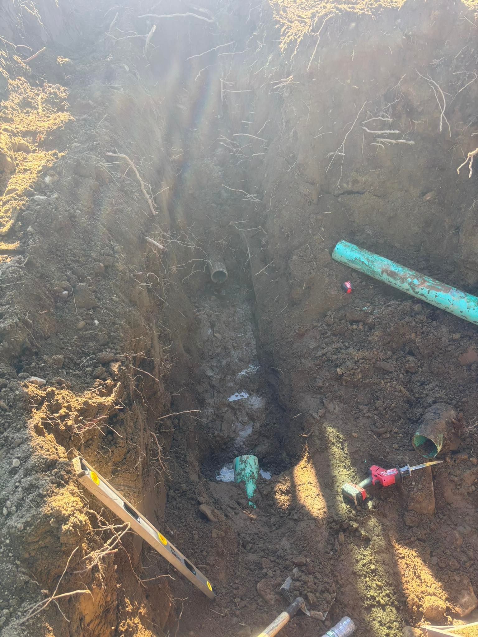 Excavation pit with dark soil, turquoise pipe, and several utility markers in bright sunlight