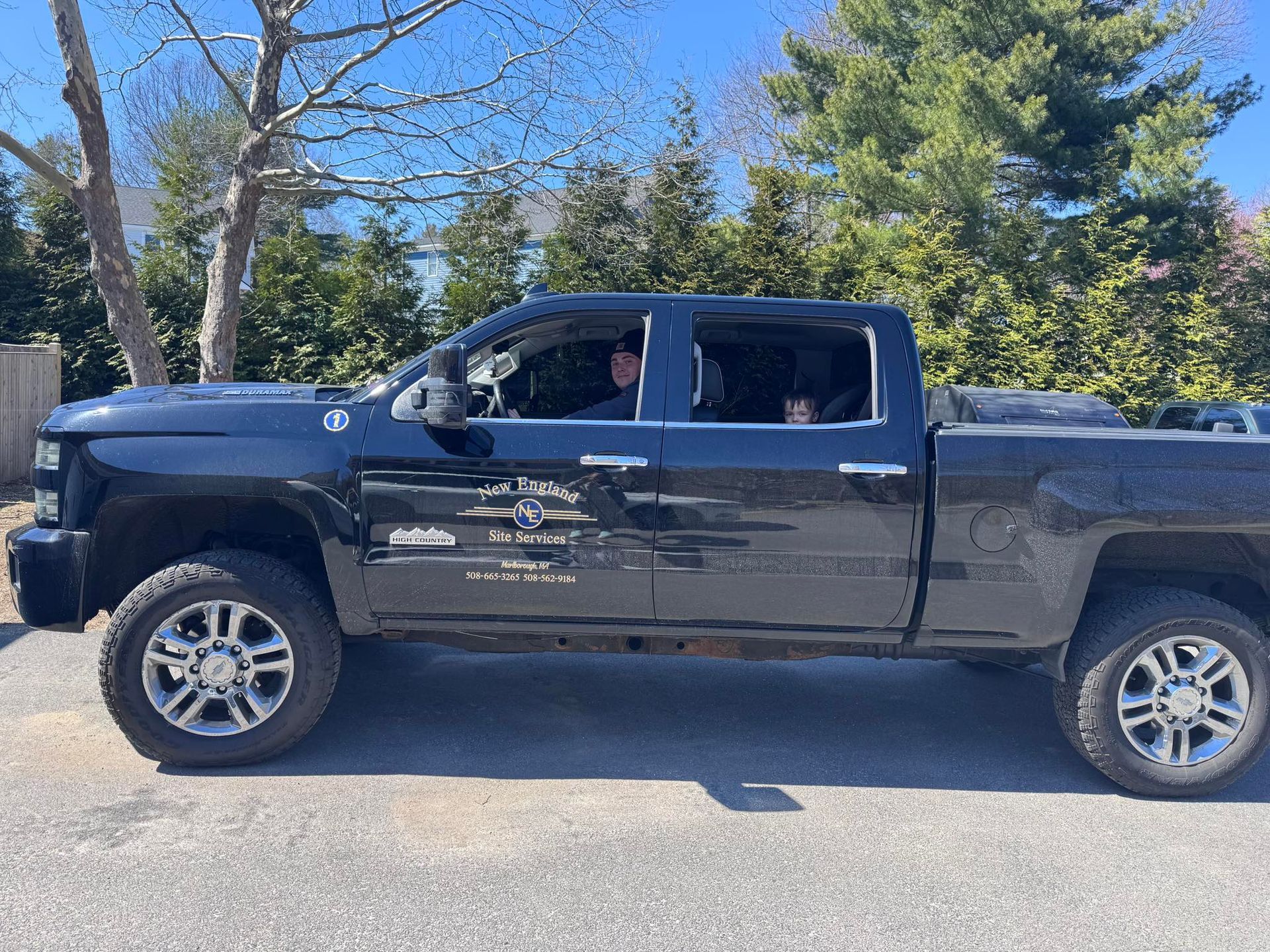 Black pickup truck parked on a road with a driver visible, trees and blue sky in the background