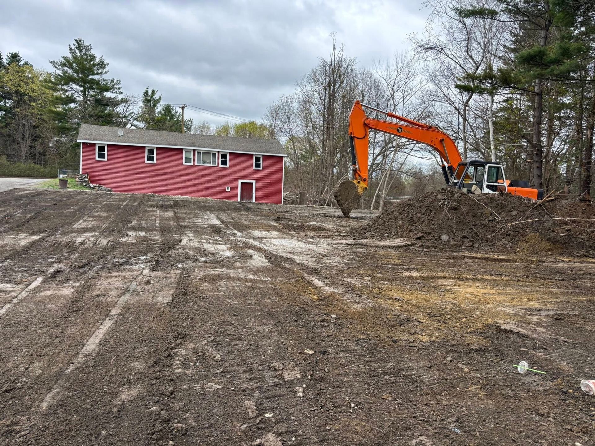 Orange excavator clearing dirt beside a red house on a muddy lot in a wooded area