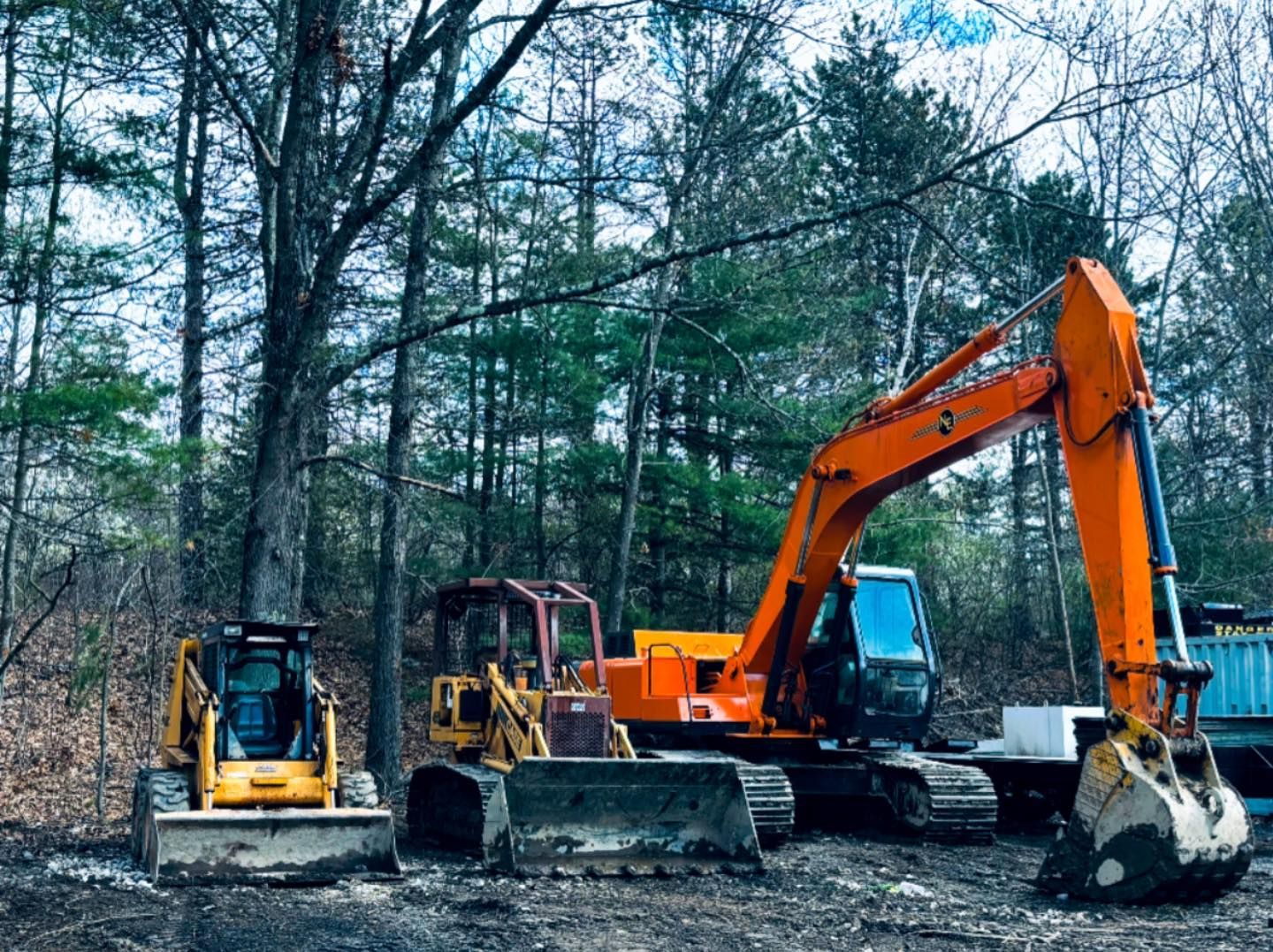 Orange excavator and two small construction machines parked on gravel in a wooded area.