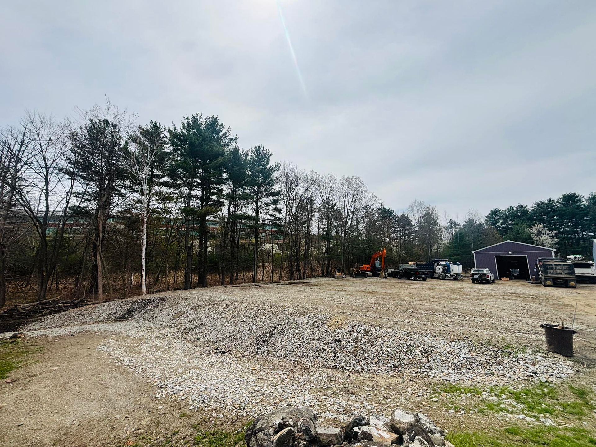 Gravel lot beside trees under cloudy sky, with a small building and parked equipment in the distance