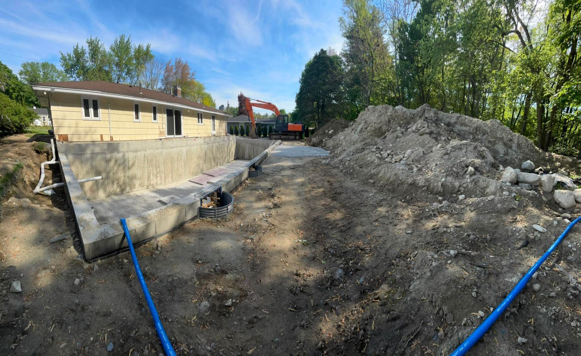 Construction site beside a beige building with dirt piles, pipes, and blue hoses under a clear sky