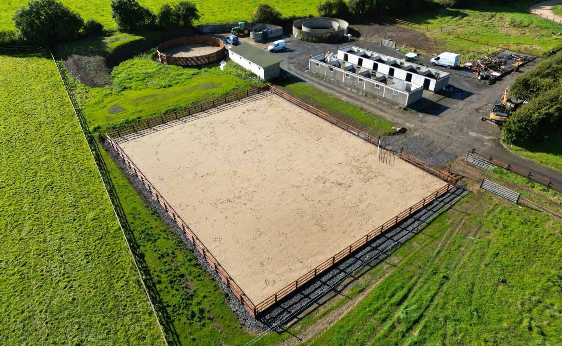 Aerial view of a fenced sand court beside green fields and farm buildings.