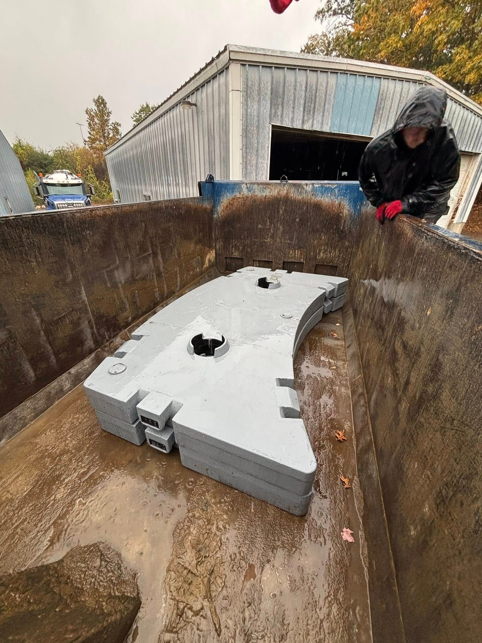 Person in rain jacket inspecting a large gray object inside a rusty metal container outdoors.
