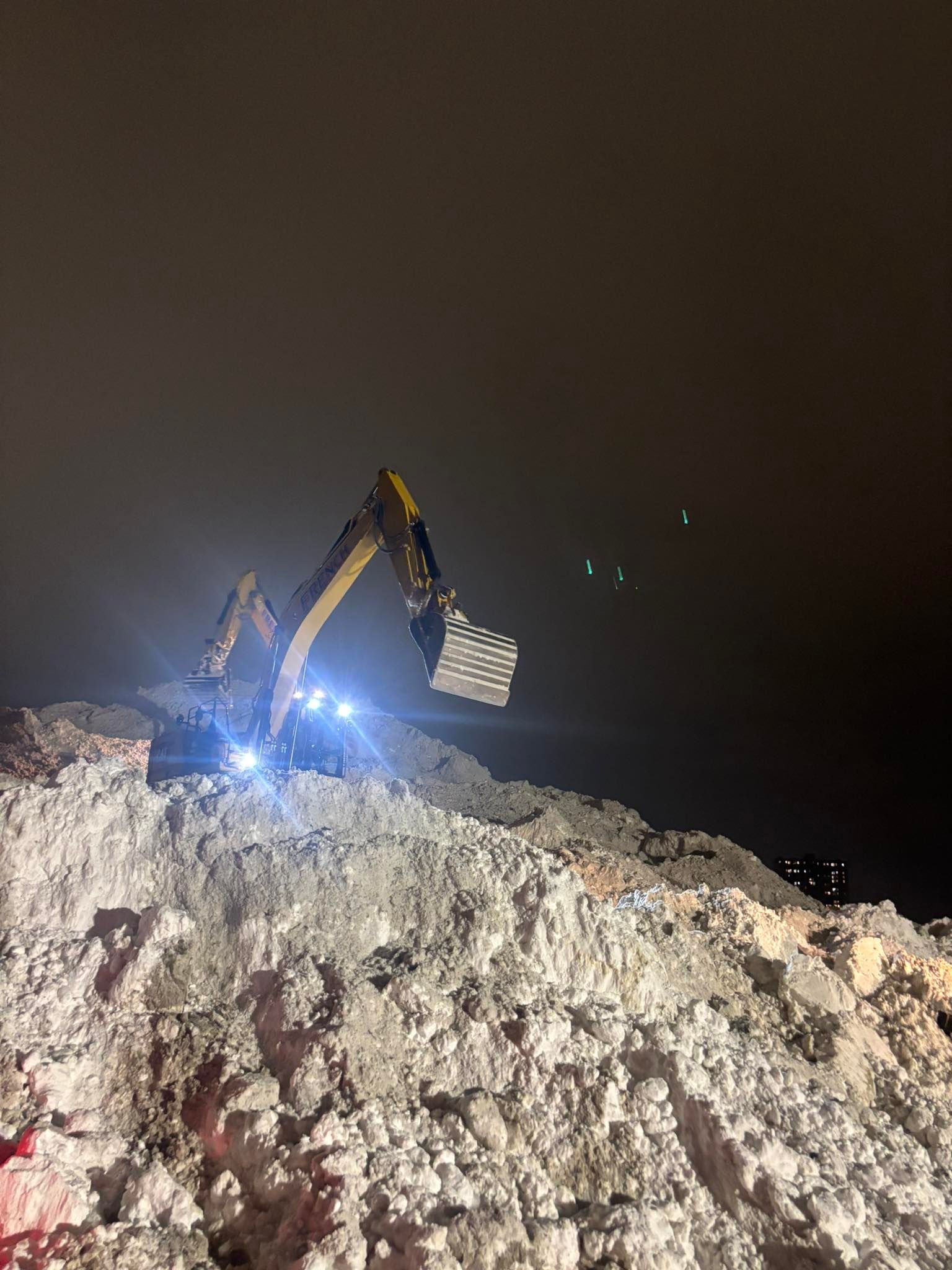 Excavator working atop a snow pile at night, lit by bright work lights under a dark sky