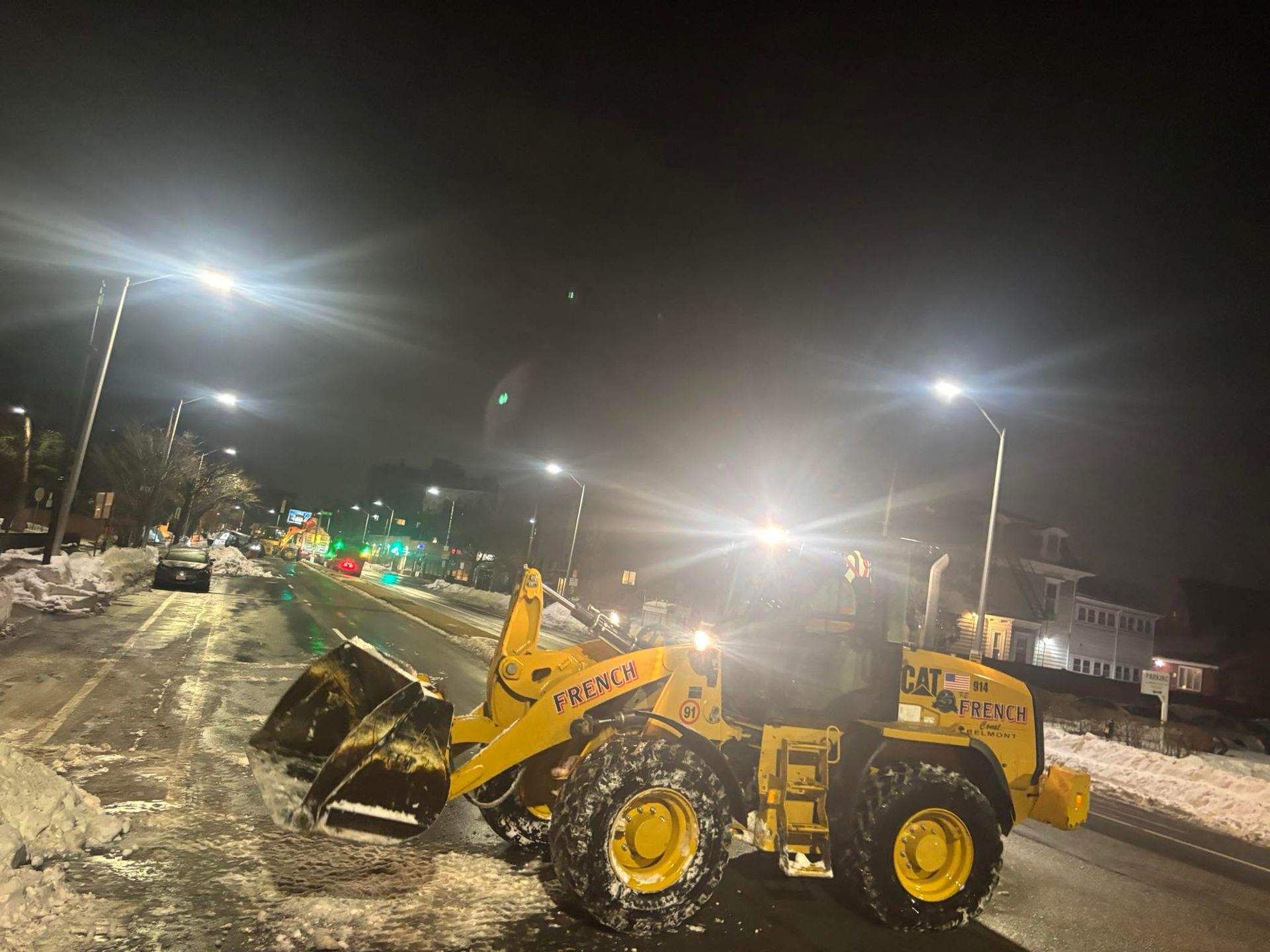 Yellow front loader plows snow on a snowy street at night under bright streetlights.