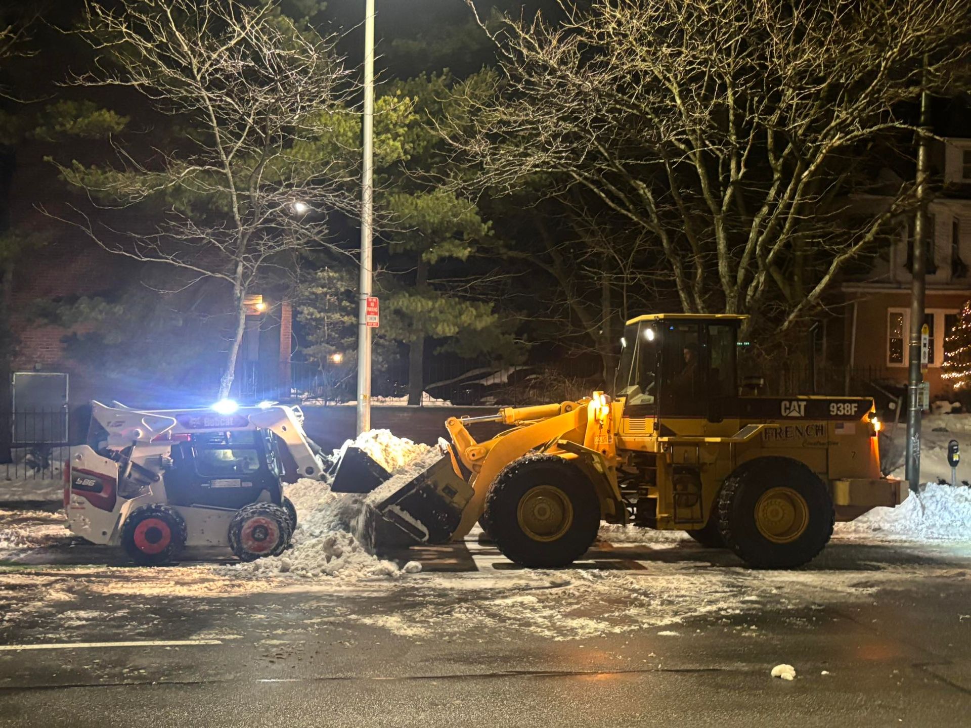Yellow front loader lifting snow beside a small snowplow on a snowy street at night, under streetlights.