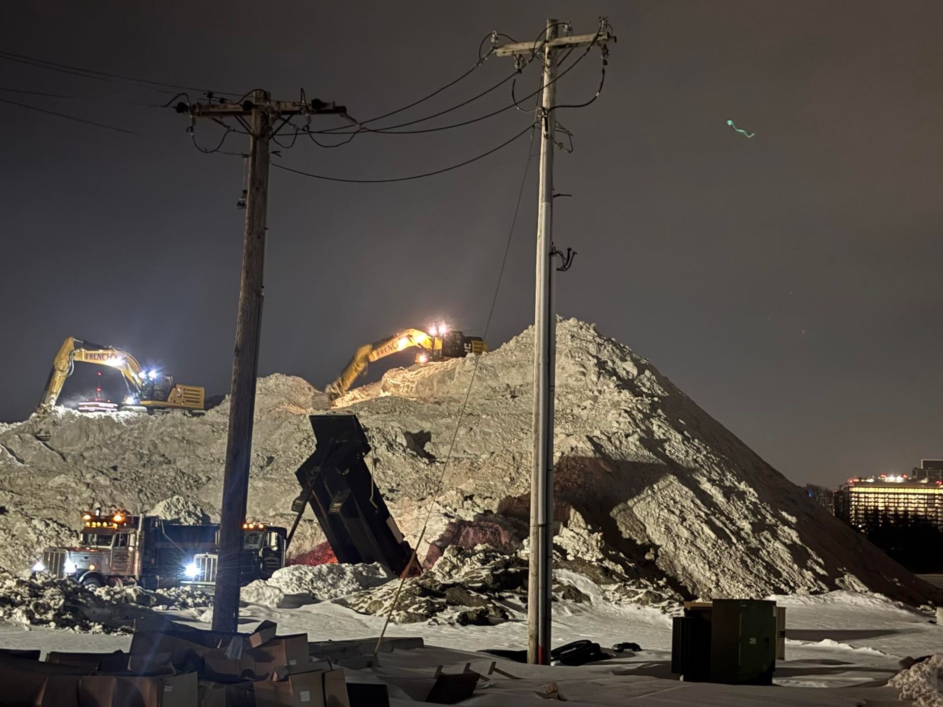 Night construction site with excavators on a rubble mound beside utility poles and power lines.