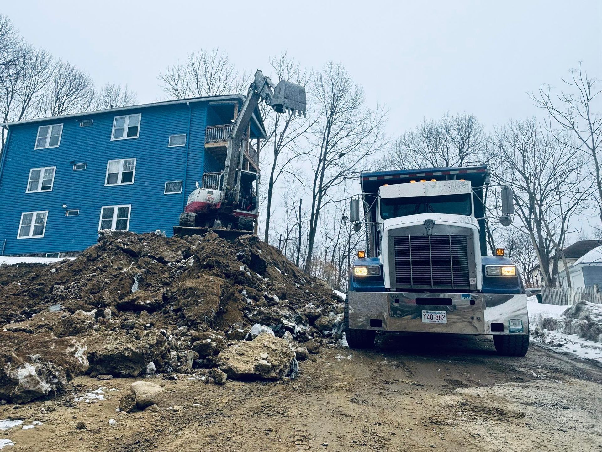 Excavator and dump truck beside a blue apartment building on a muddy construction site