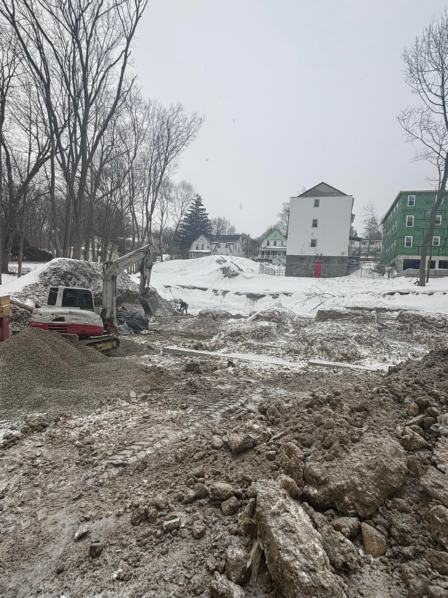Snowy muddy construction site with excavated ground, bare trees, and houses in the background