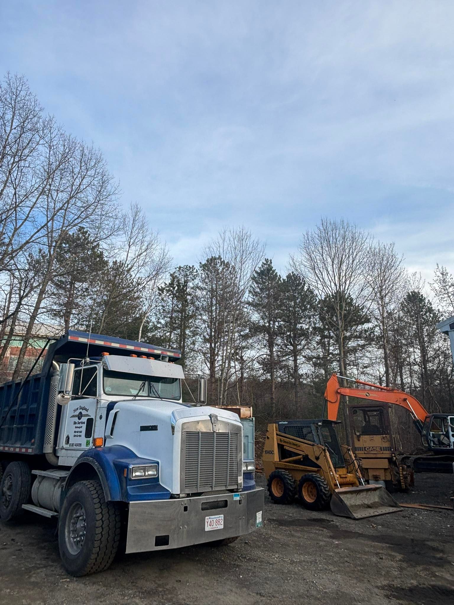 Blue dump truck and orange excavator in a dirt lot under a cloudy sky