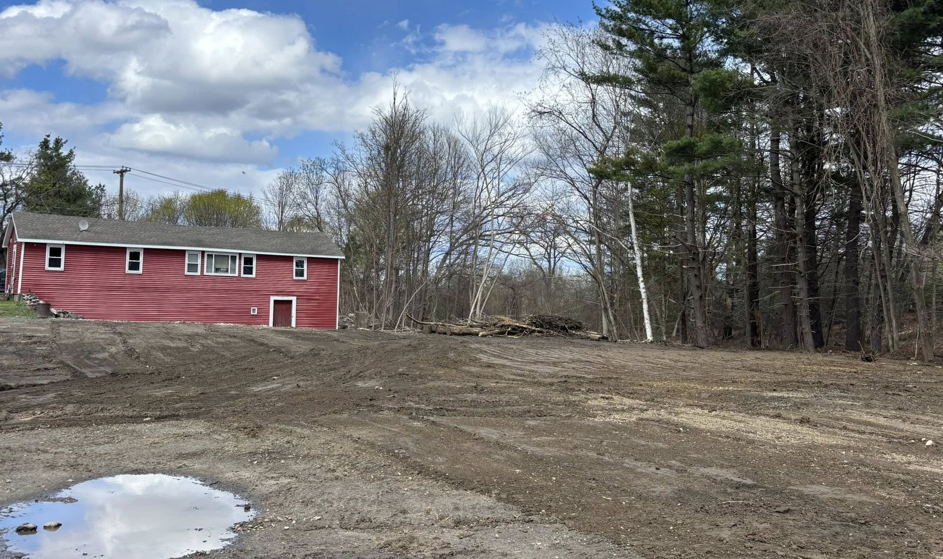 Red house beside a muddy clearing and forest under a cloudy sky