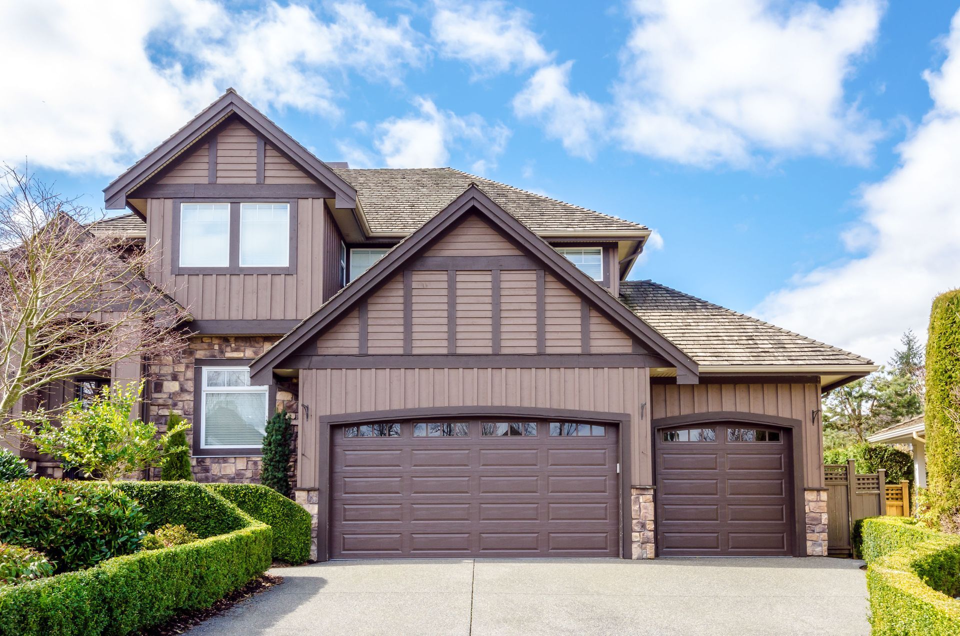 Brown two-story house with a two-car garage under a blue sky with fluffy clouds.