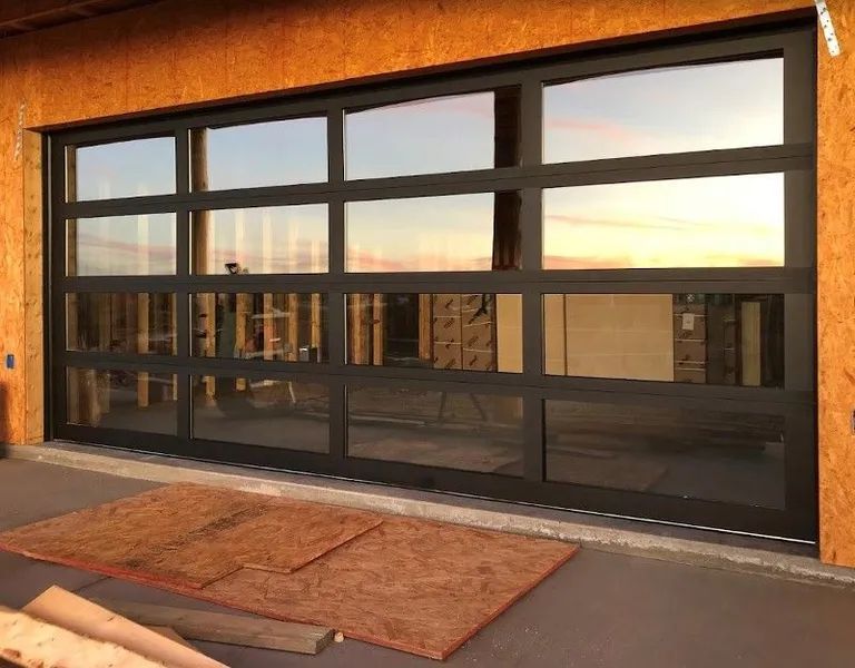 Black-framed glass garage door reflecting a sunset. Door is on a wooden building with a brown mat on the floor.