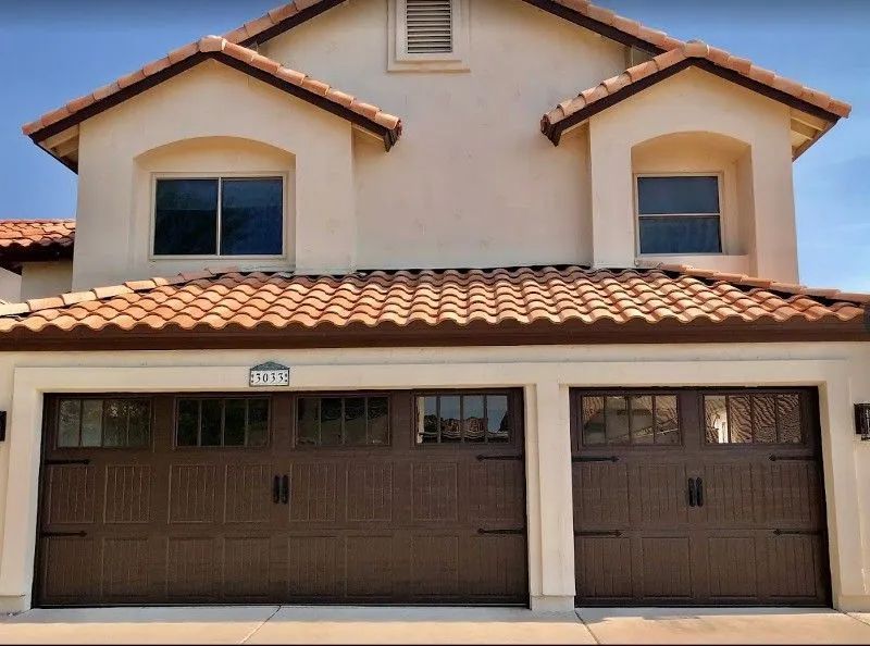 Brown garage doors on a beige house with a tiled roof and small windows above.