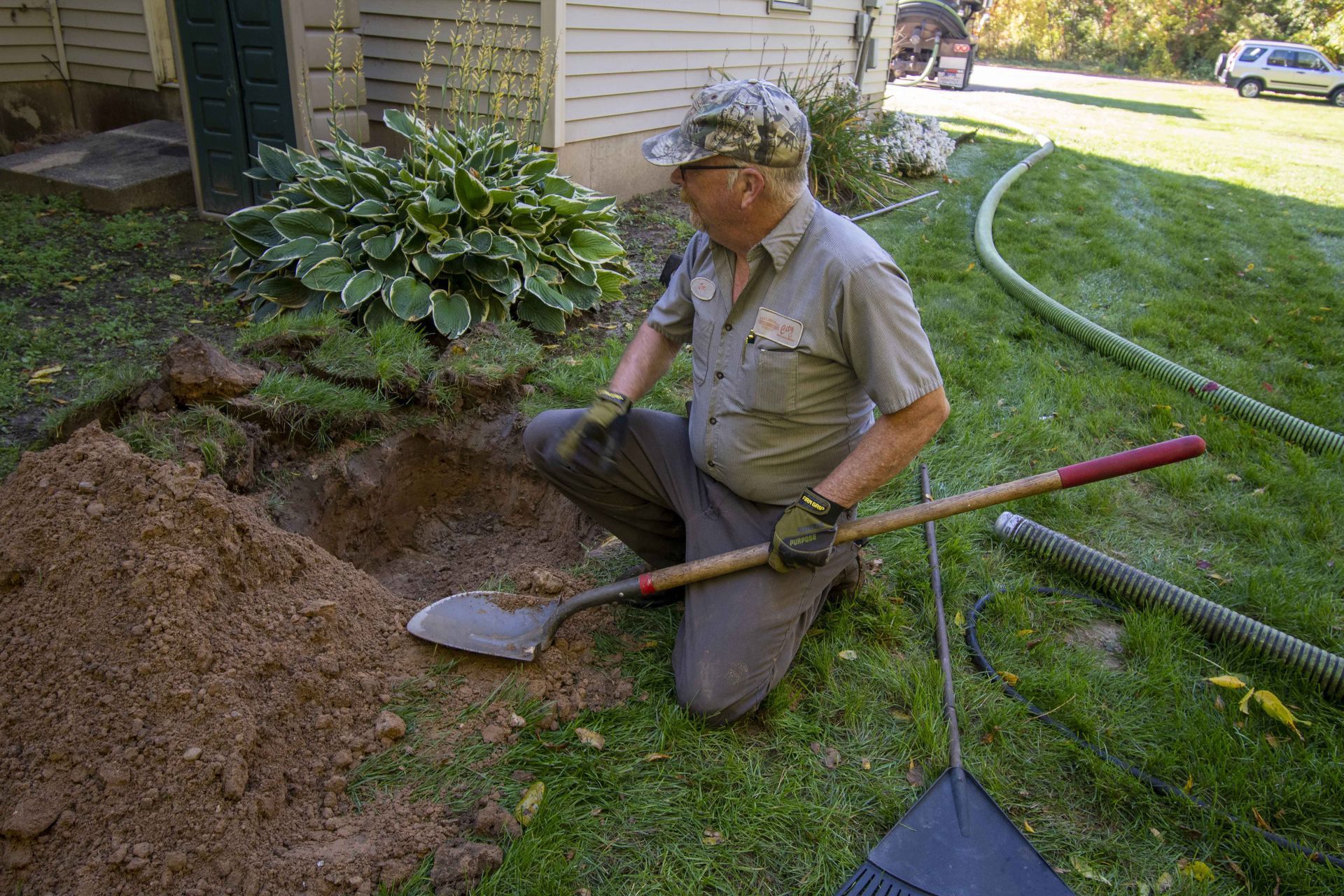 Man kneeling, digging a hole in a grassy yard near a building. He wears gloves and work attire.