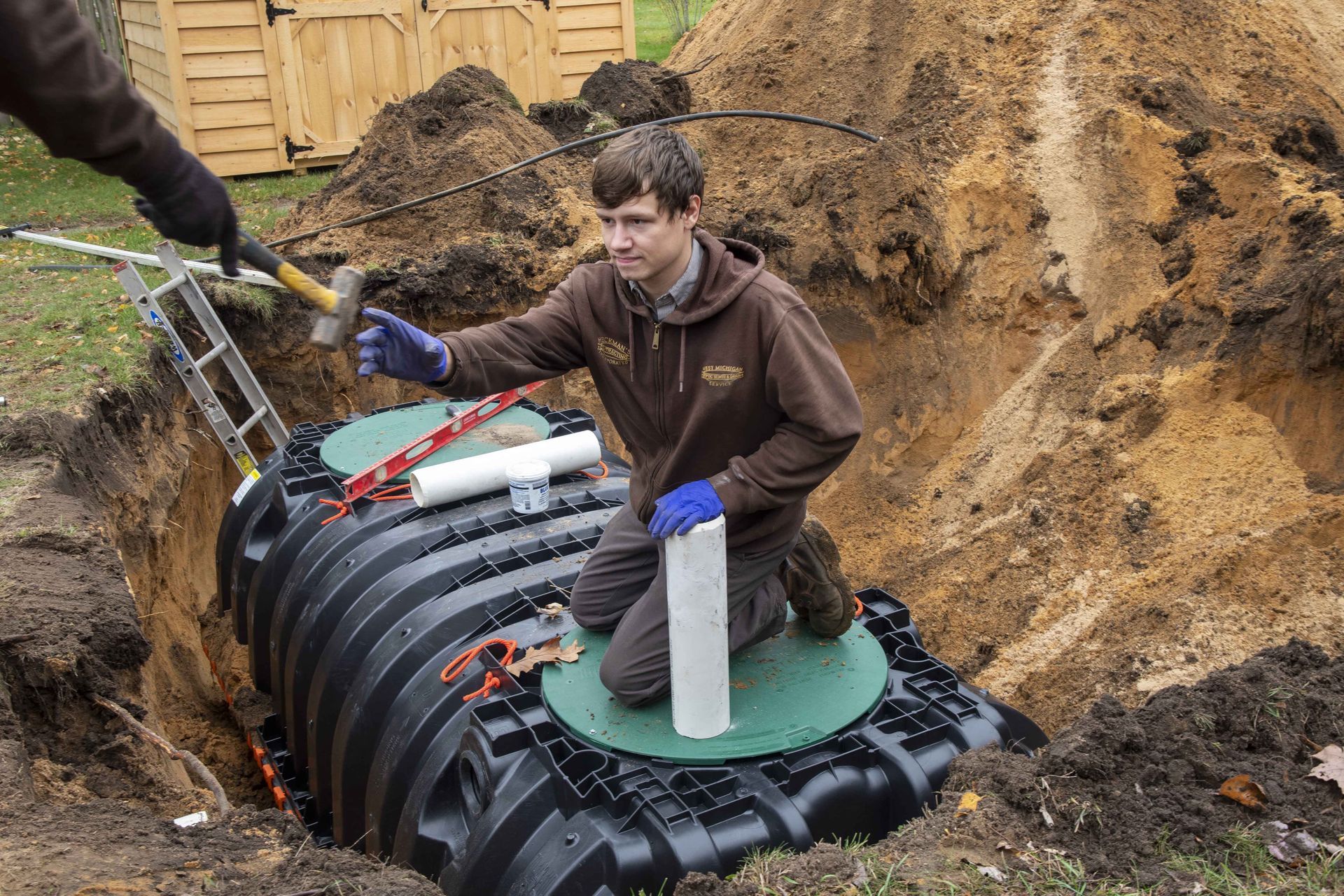 Man installing septic tank in an outdoor setting, kneeling inside excavated area. Another person assisting.