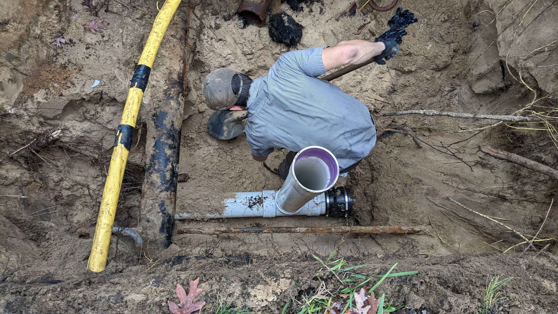 Man in trench repairing pipe, wearing gloves and hat. Dirt, yellow measuring tape, and PVC pipe visible.