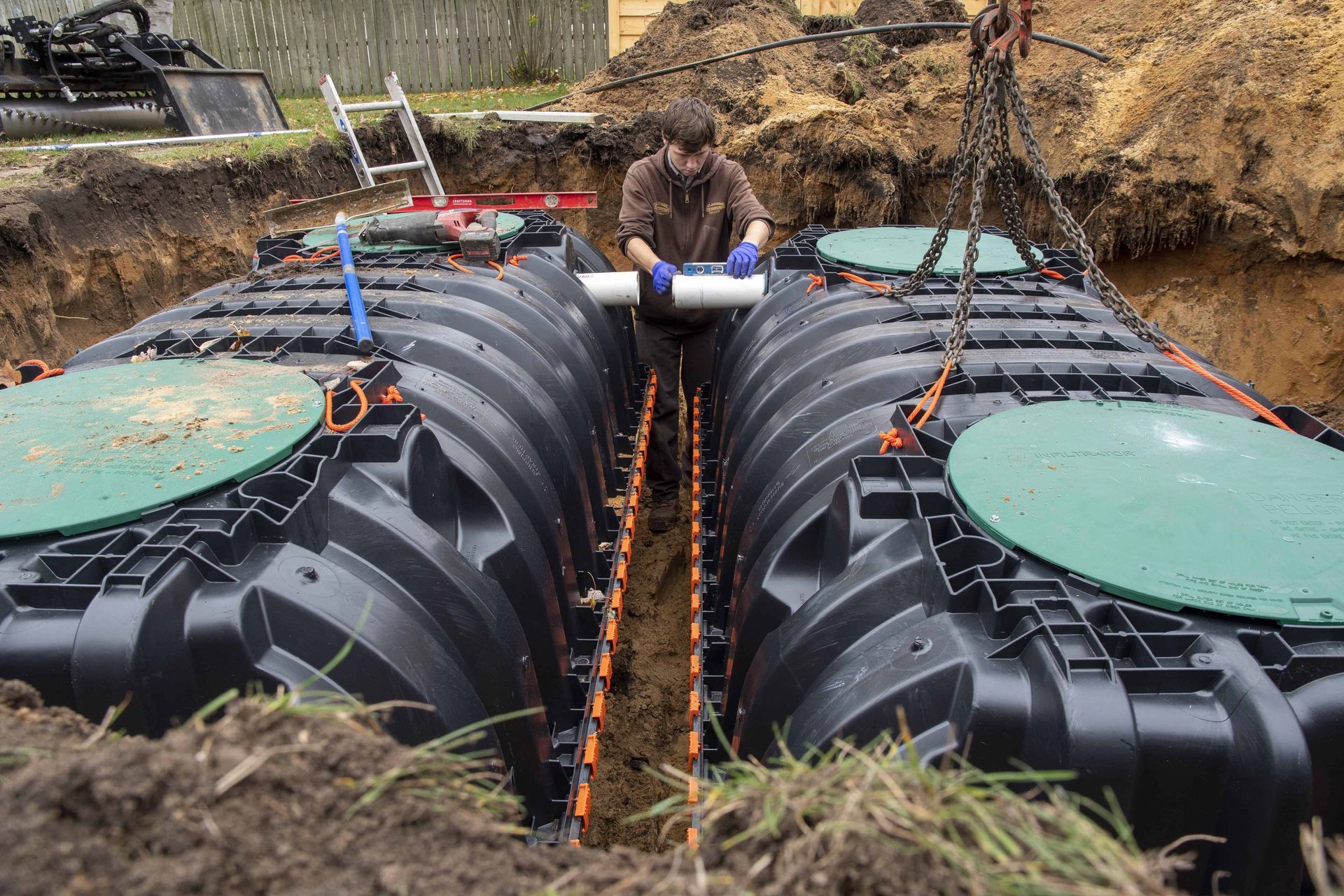 A person installing pipes between two large, black septic tanks in an excavated area.