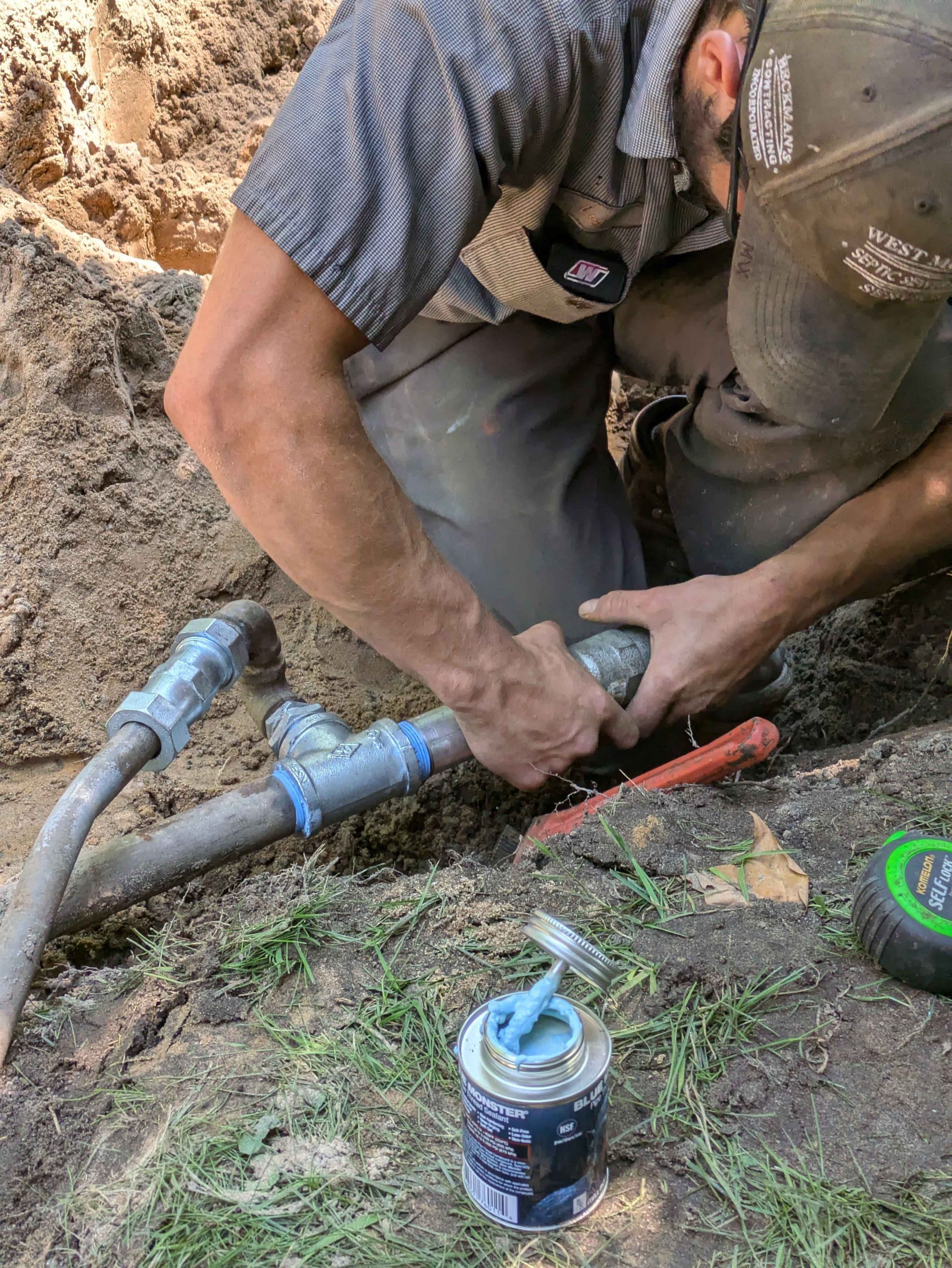 Plumber kneels in a trench, connecting pipes with glue; blue glue container visible.