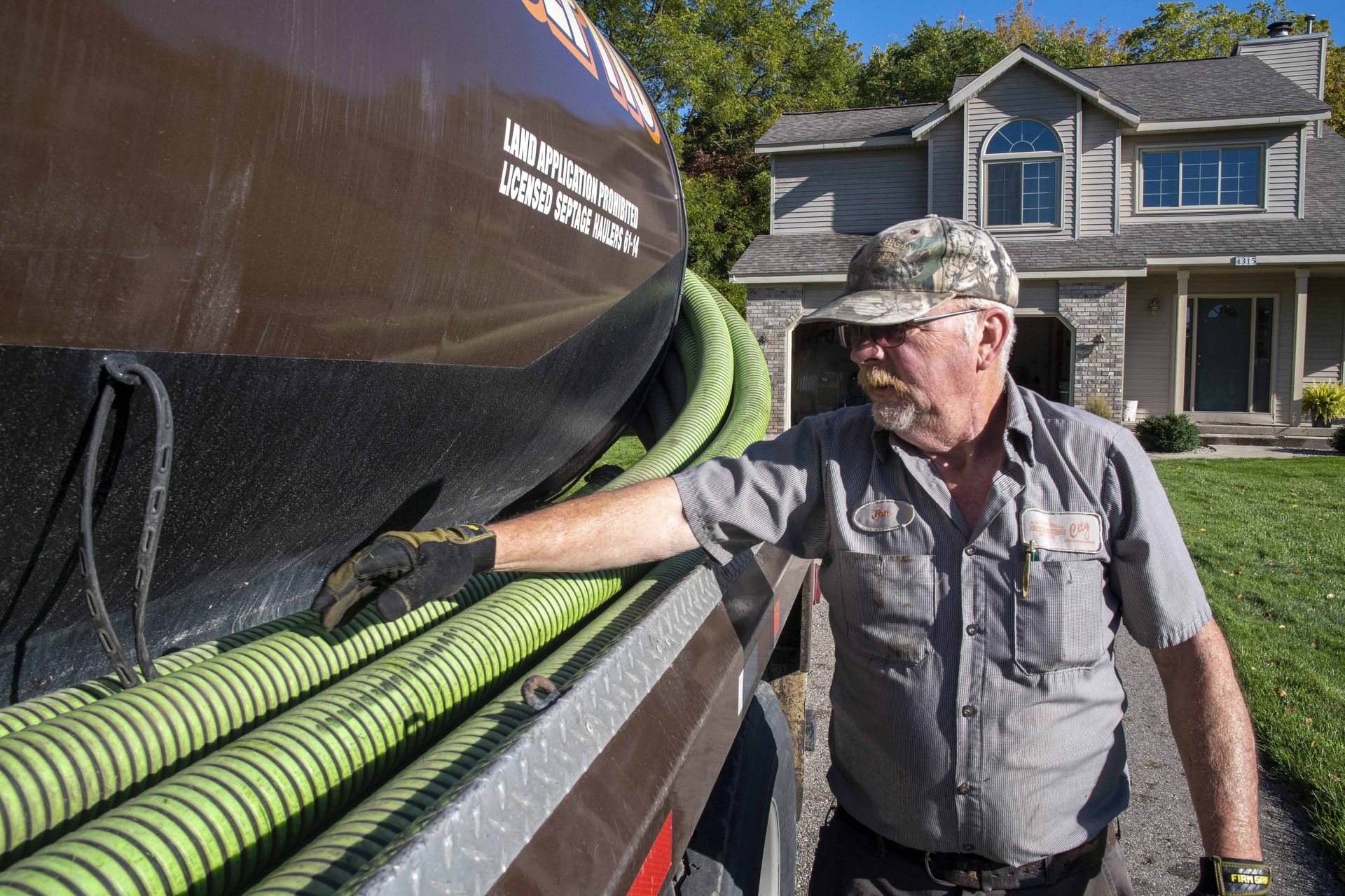 Man in work clothes beside a brown septic tank truck, examining green hoses, in front of a house.