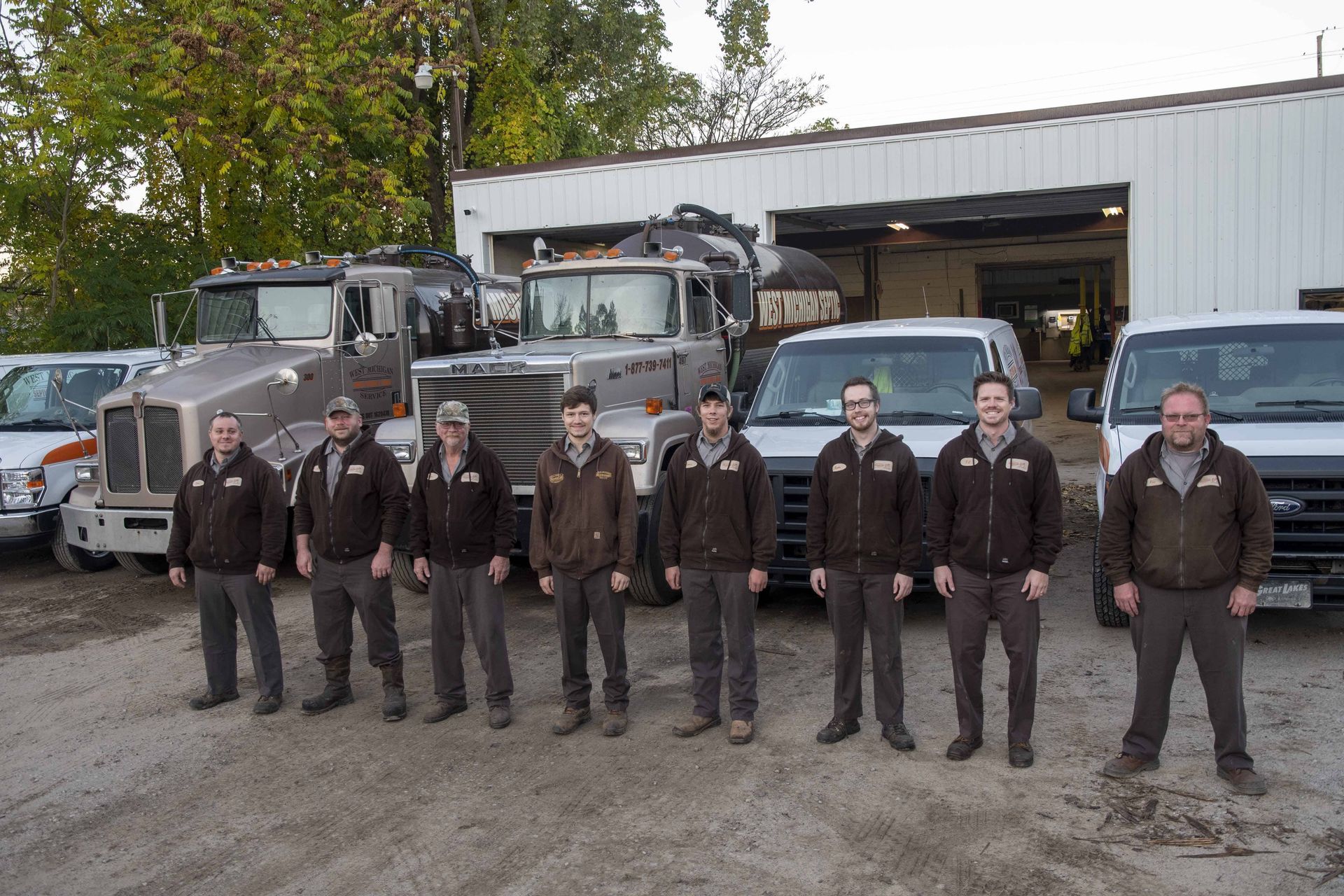 Group of nine people in front of tow trucks and garage. Men are wearing matching jackets.