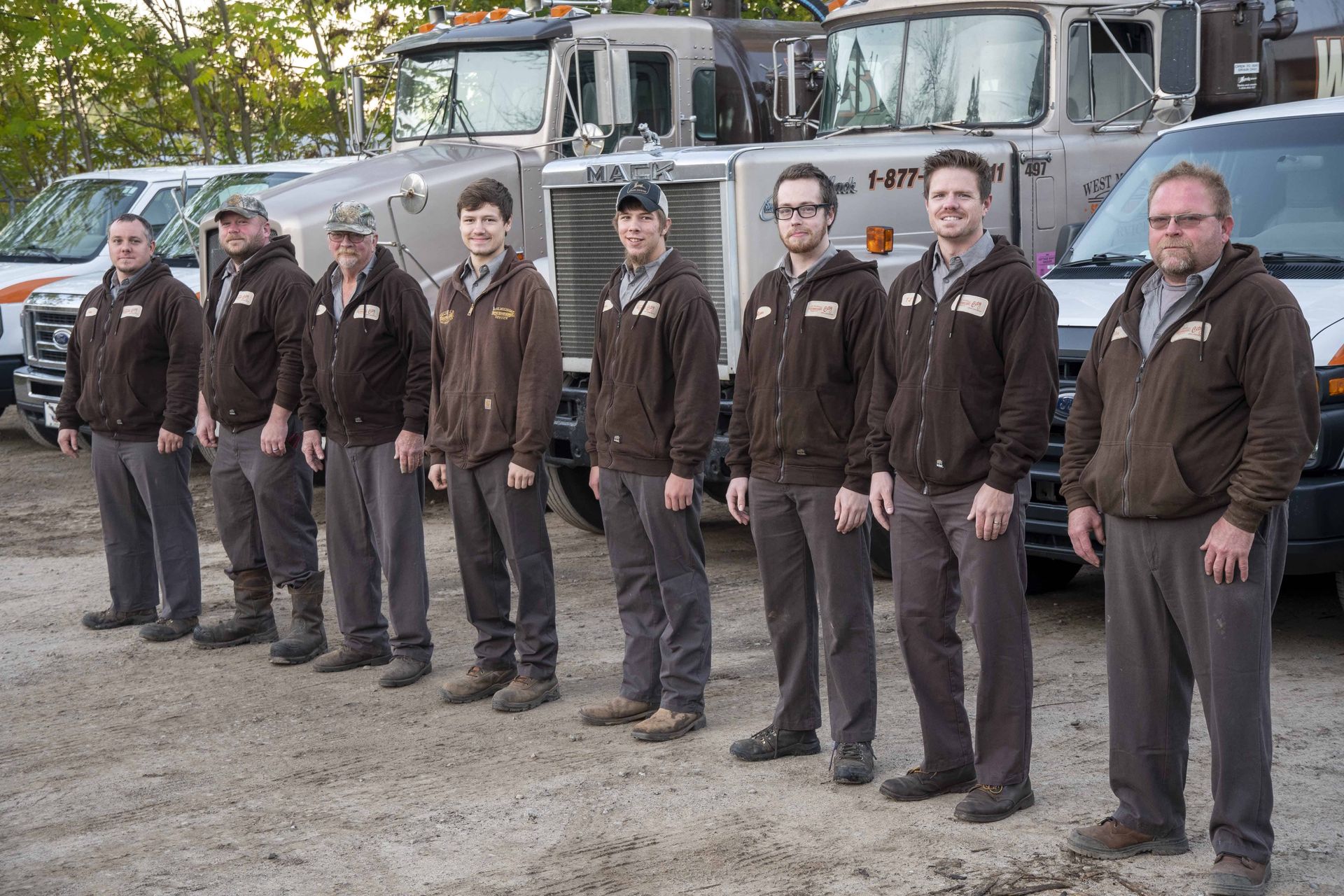 Eight men in brown jackets and grey pants stand in front of utility trucks, likely a work crew.
