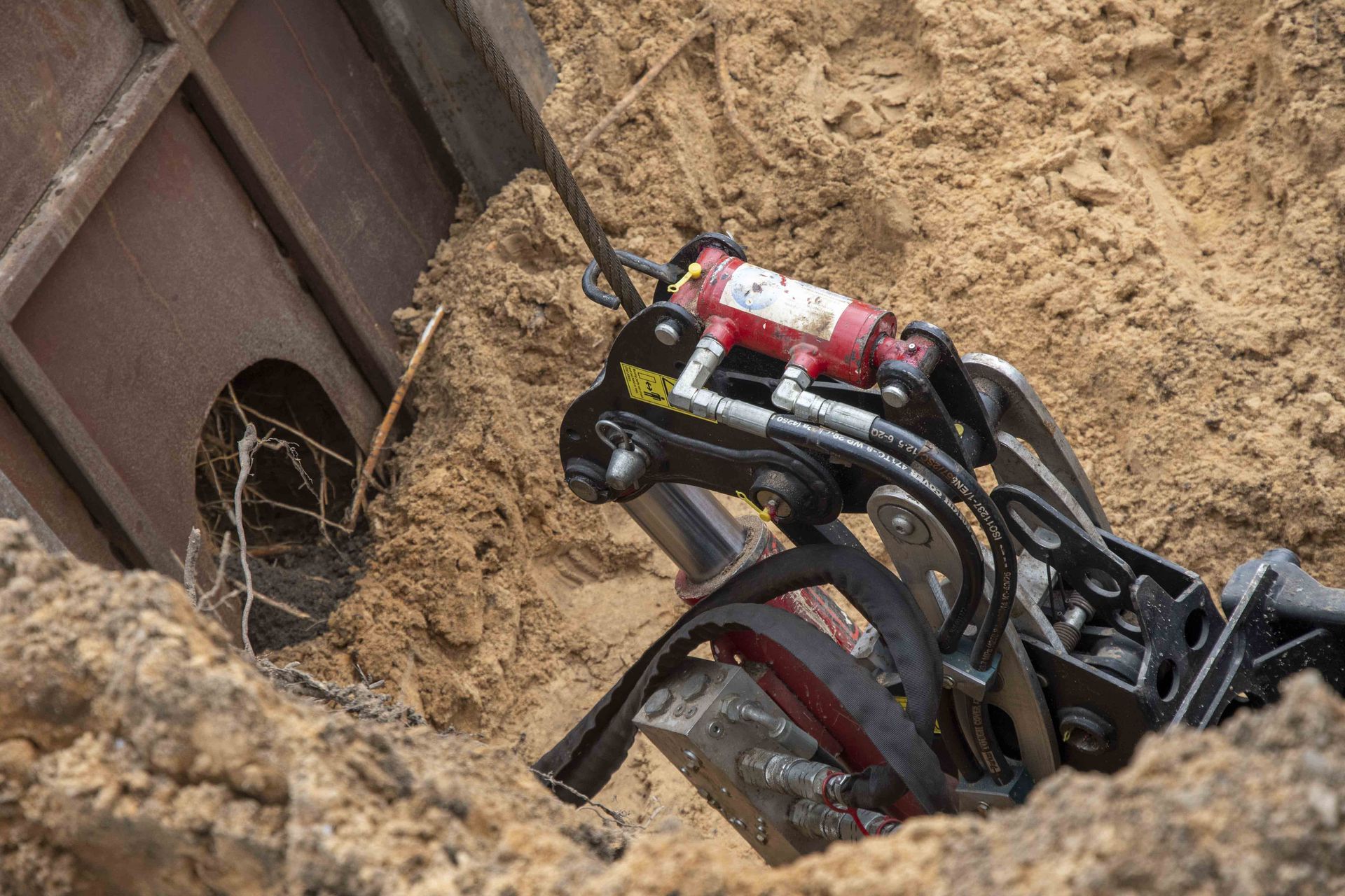 A hydraulic device digging in the sand, near a dark wooden structure.
