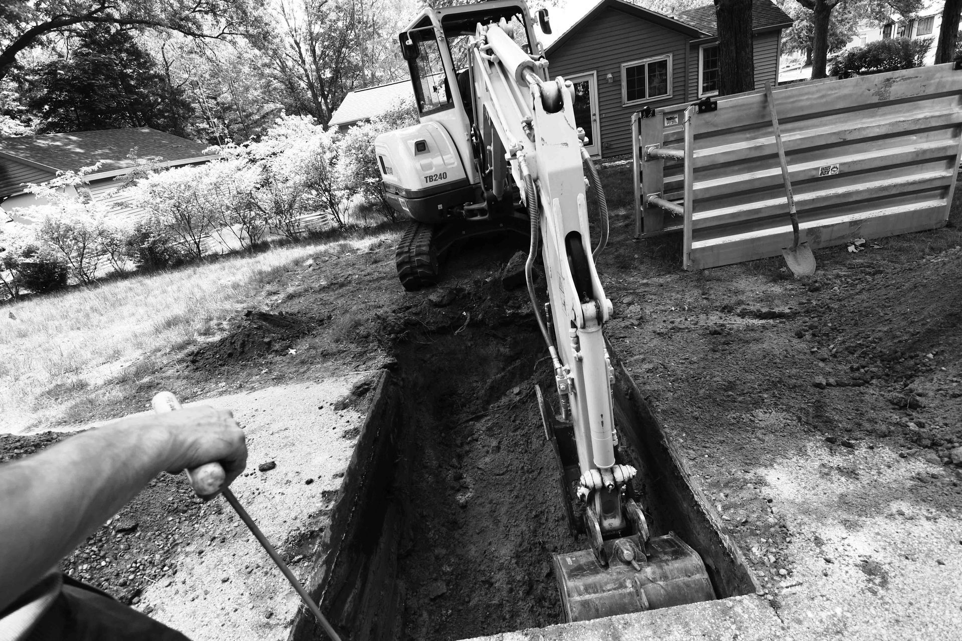 An excavator digs a trench in a yard; a person operates controls.