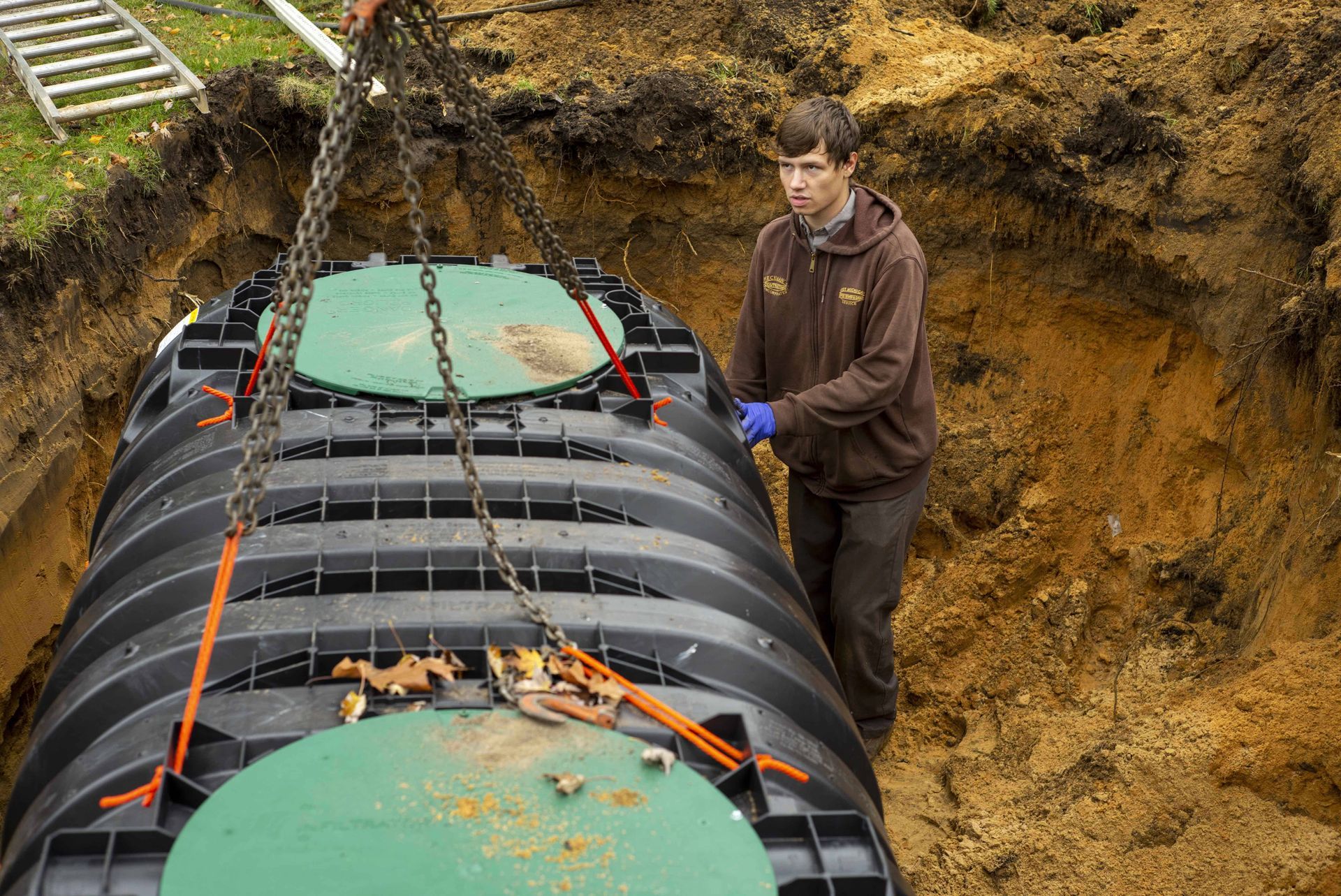 Man in brown hoodie guides a large black septic tank being lowered into a dirt excavation.