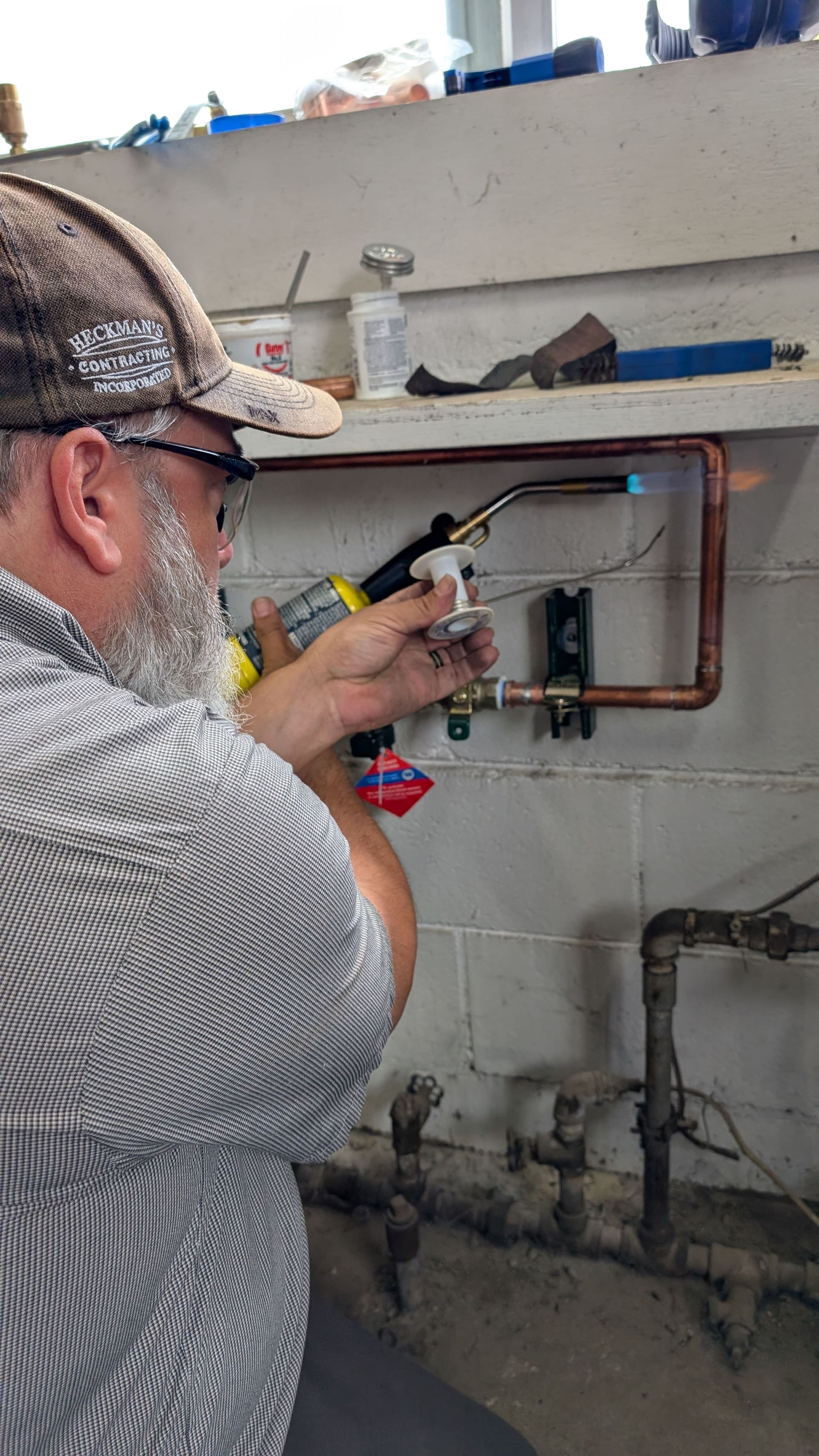 Man soldering copper pipes near a wall. He's wearing a hat and glasses. The setting is likely a utility room.