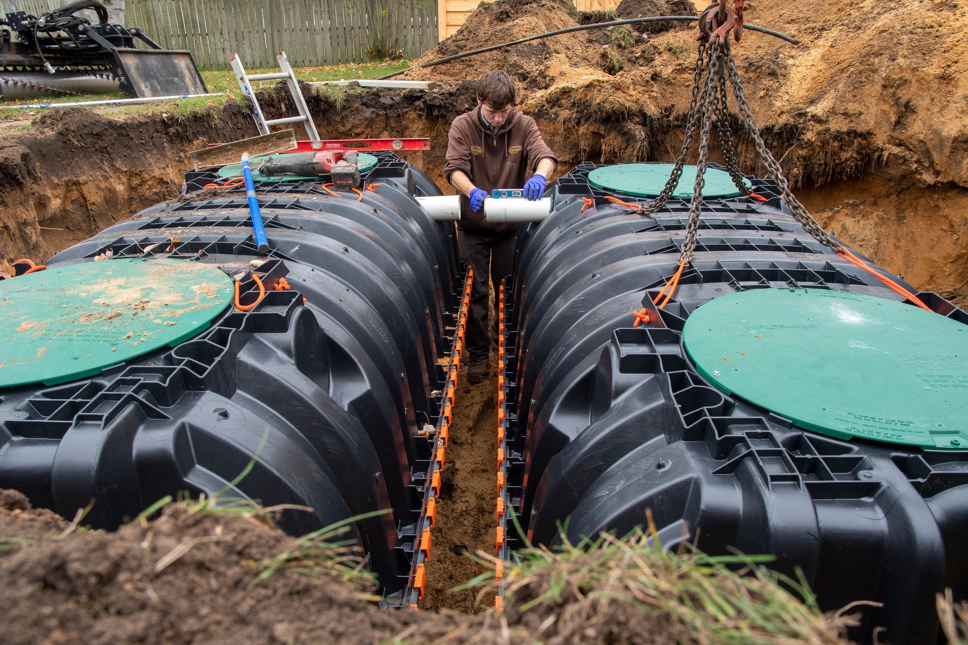 Man connecting pipes to two black septic tanks in an excavated trench. Green lids, outdoor setting.