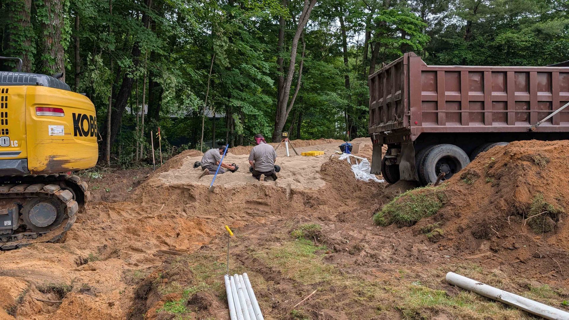 Construction site: excavator, dump truck, workers digging in dirt, pipes.