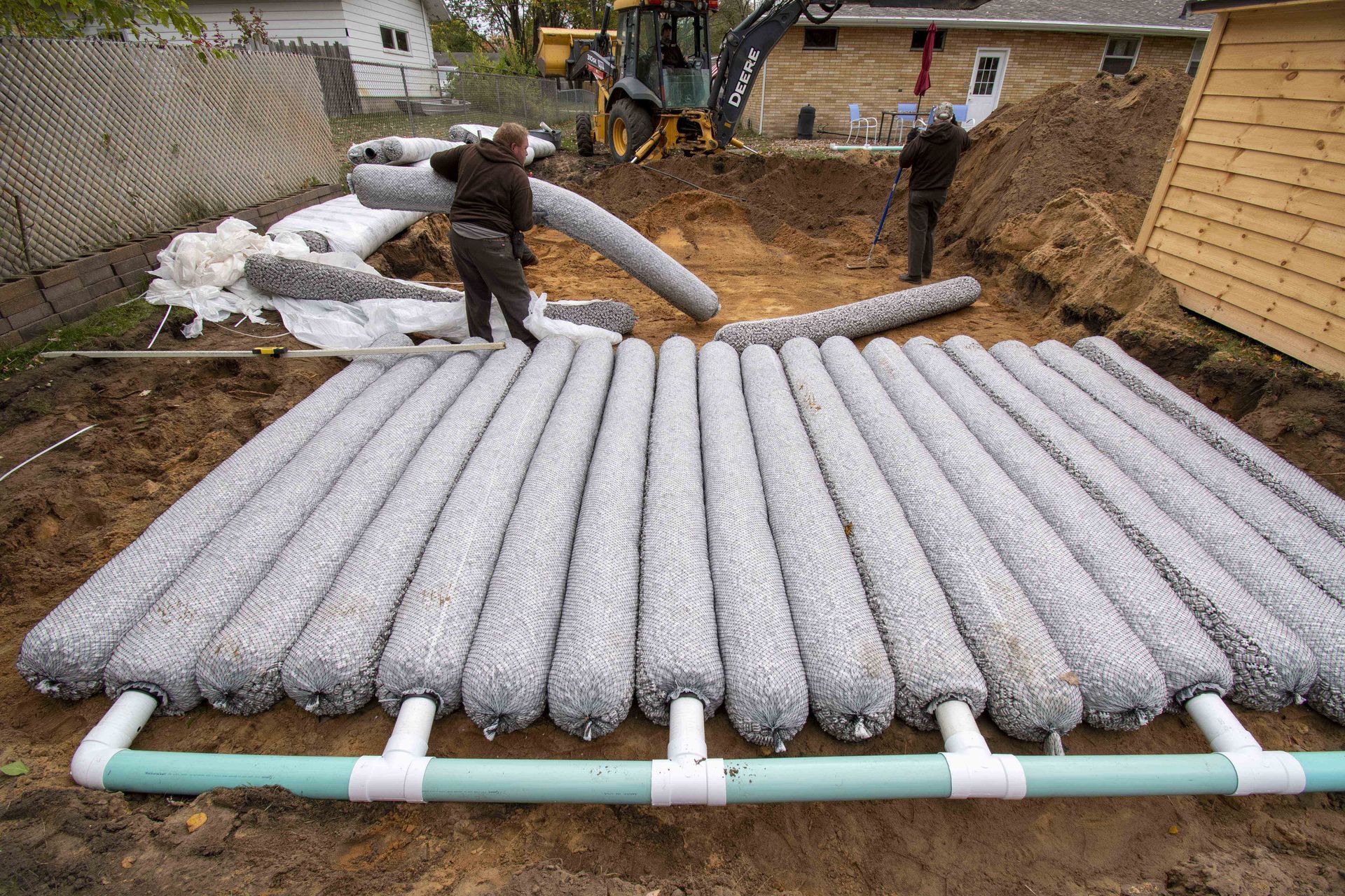 Workers installing a grey gravel-filled drainage system with green pipes in a residential yard.