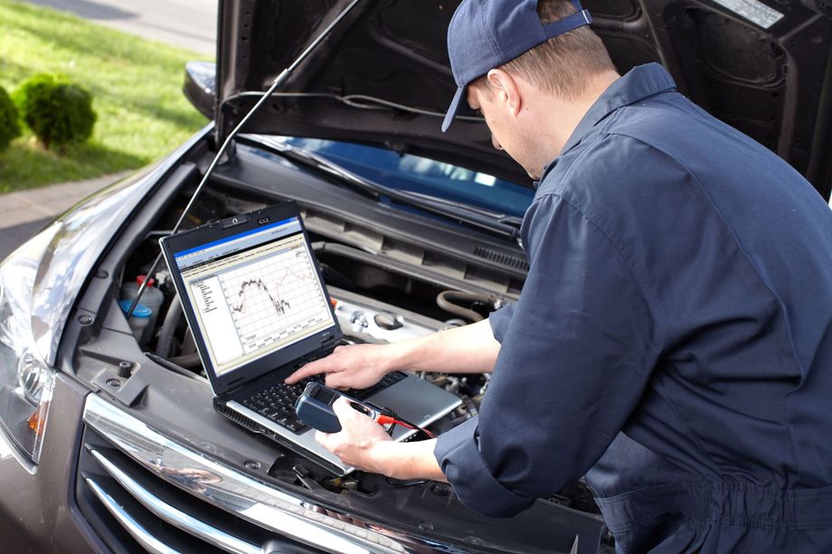 Mechanic in blue uniform uses laptop to diagnose car engine with hood open.